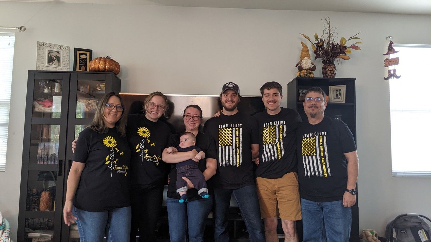 Family poses in front of a wall with black cabinets and decorations. People are wearing matching shirts.