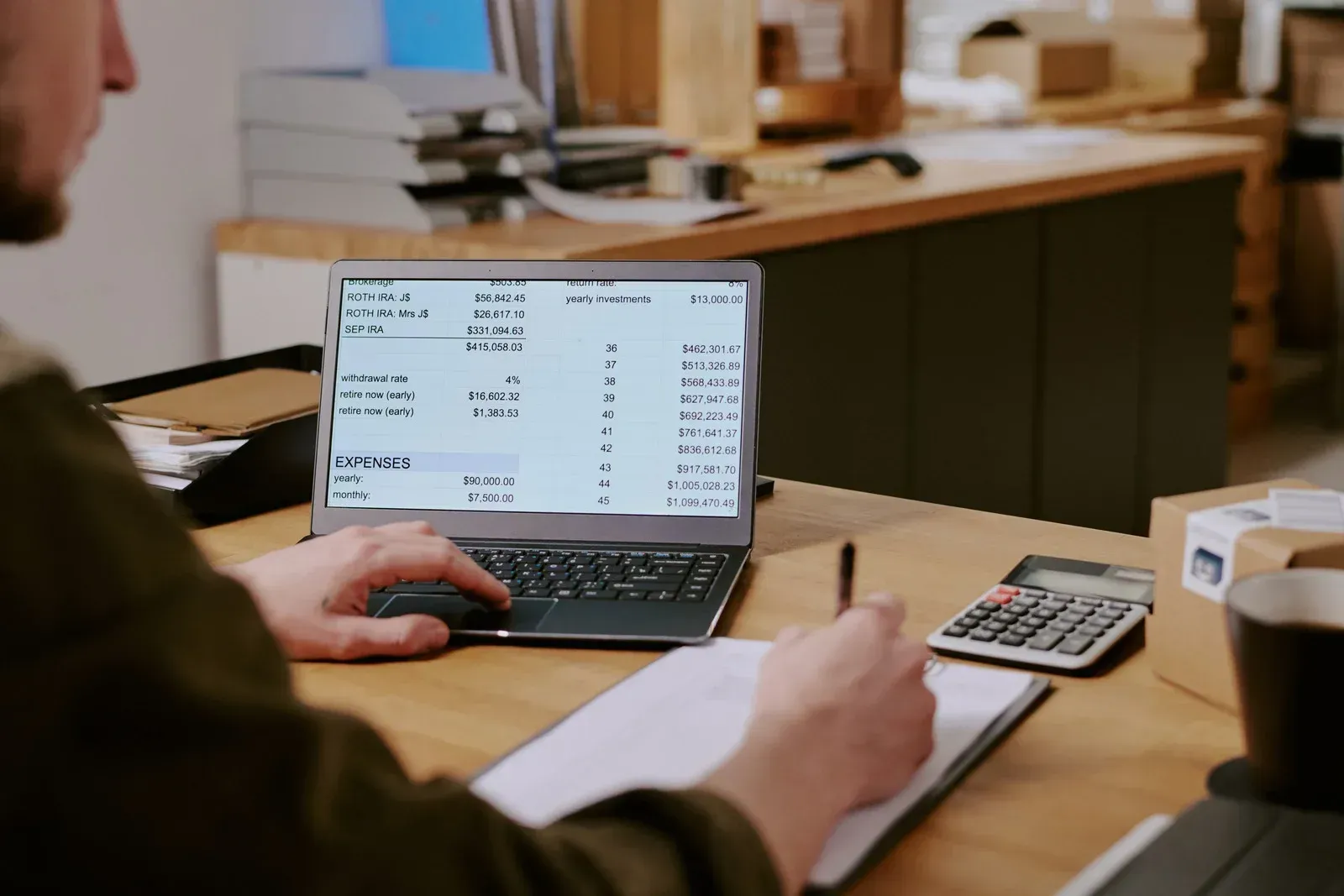 Person working on laptop at a desk, looking at numbers, and writing on a notepad with calculator nearby.