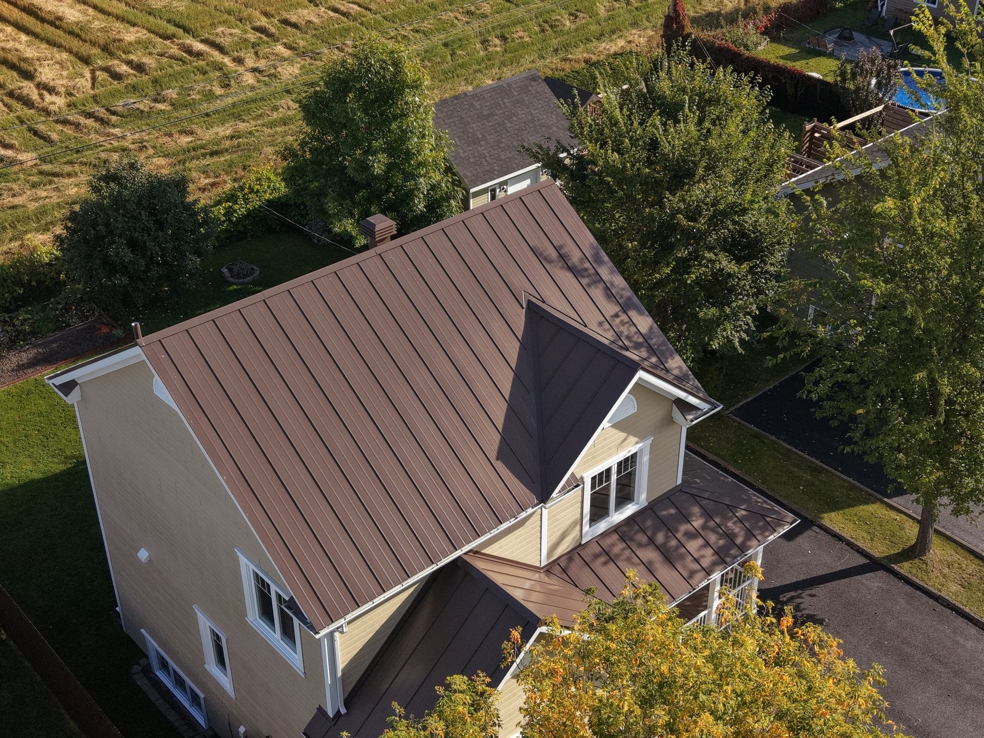 Vue aérienne d'une maison au toit brun et au bardage beige, entourée d'arbres et d'une allée pavée.