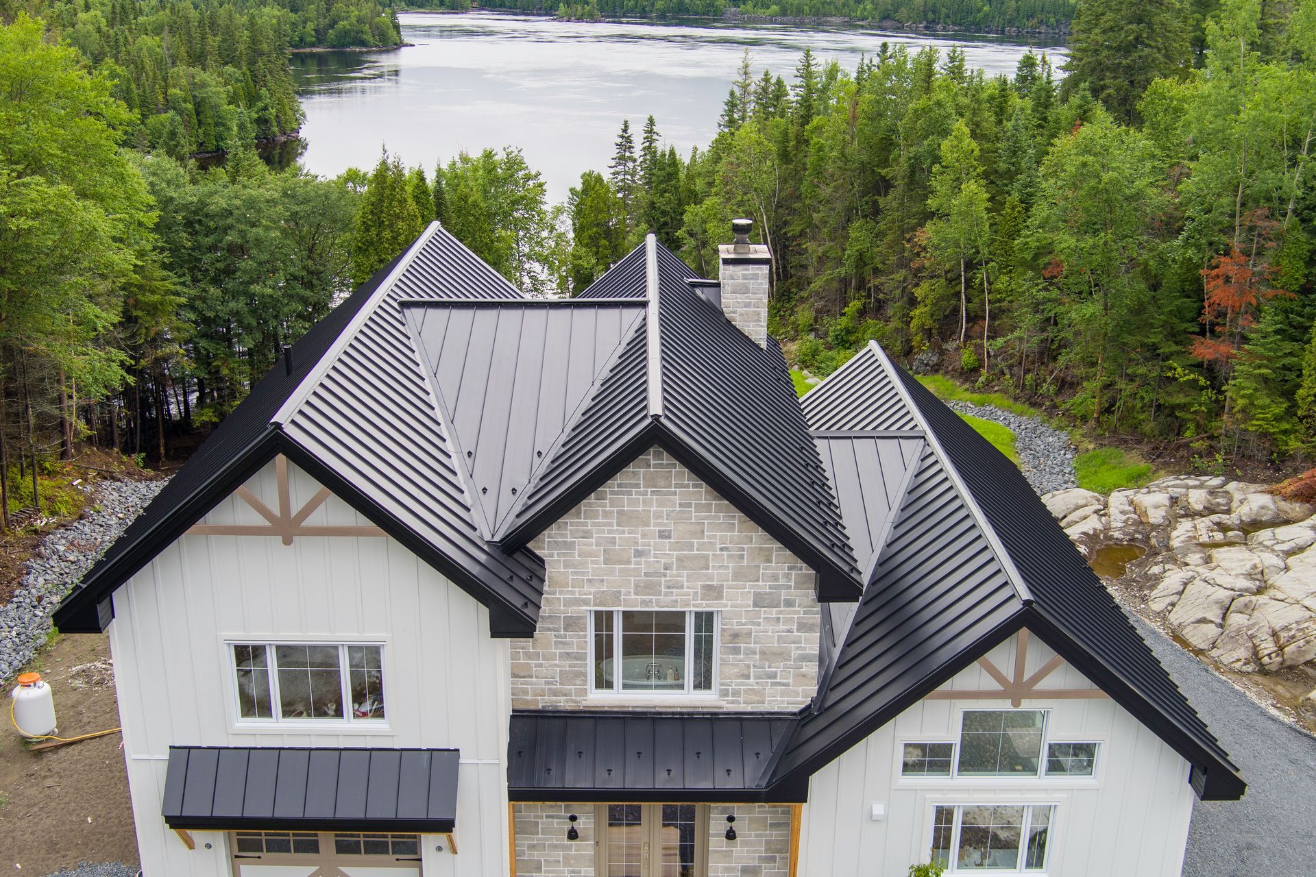 Vue aérienne d'une grande maison au toit gris, située au bord d'un lac et entourée d'arbres.