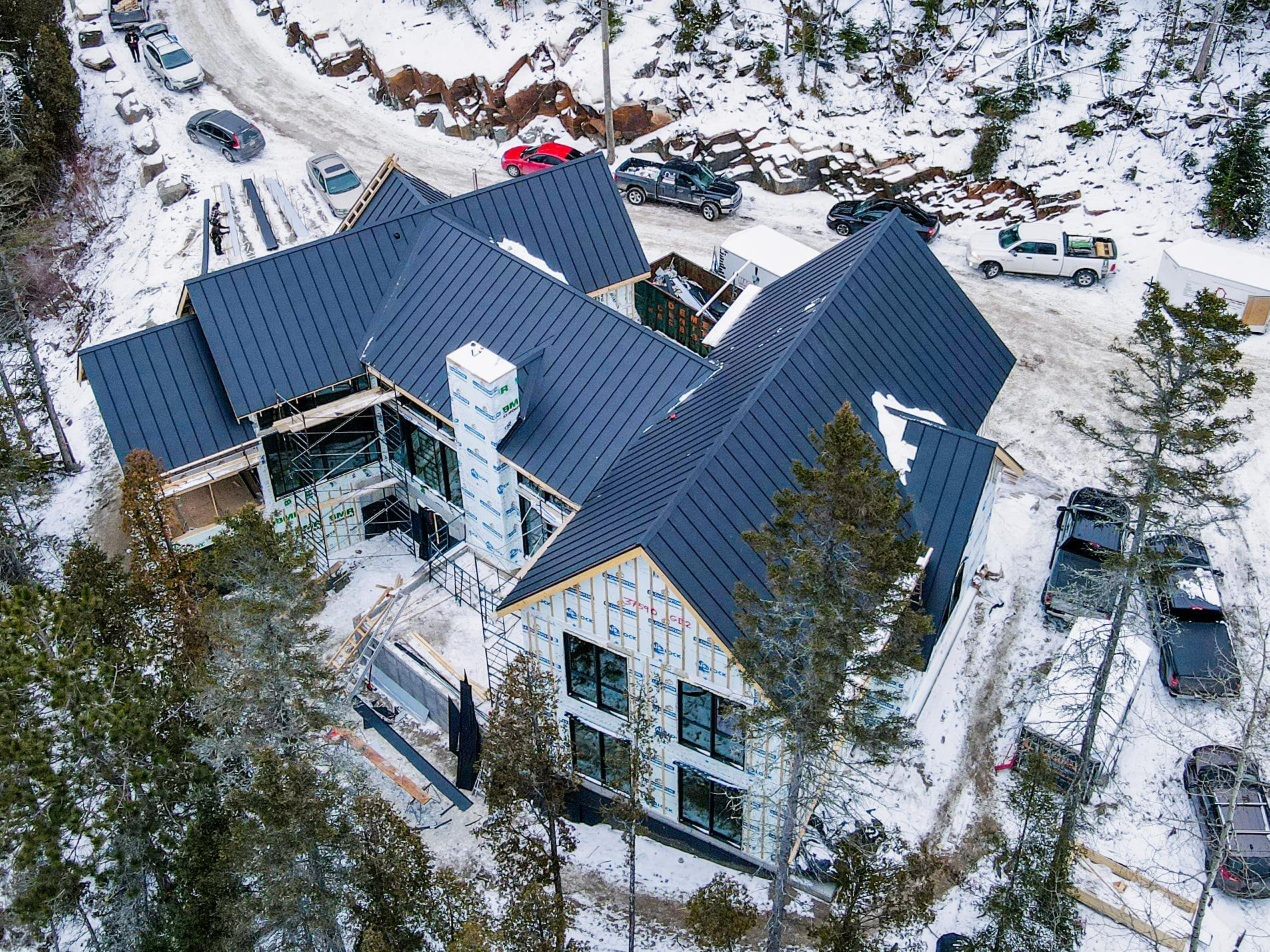 Vue aérienne d'une grande maison en construction, au toit sombre, dans un environnement enneigé, entourée d'arbres et de véhicules.