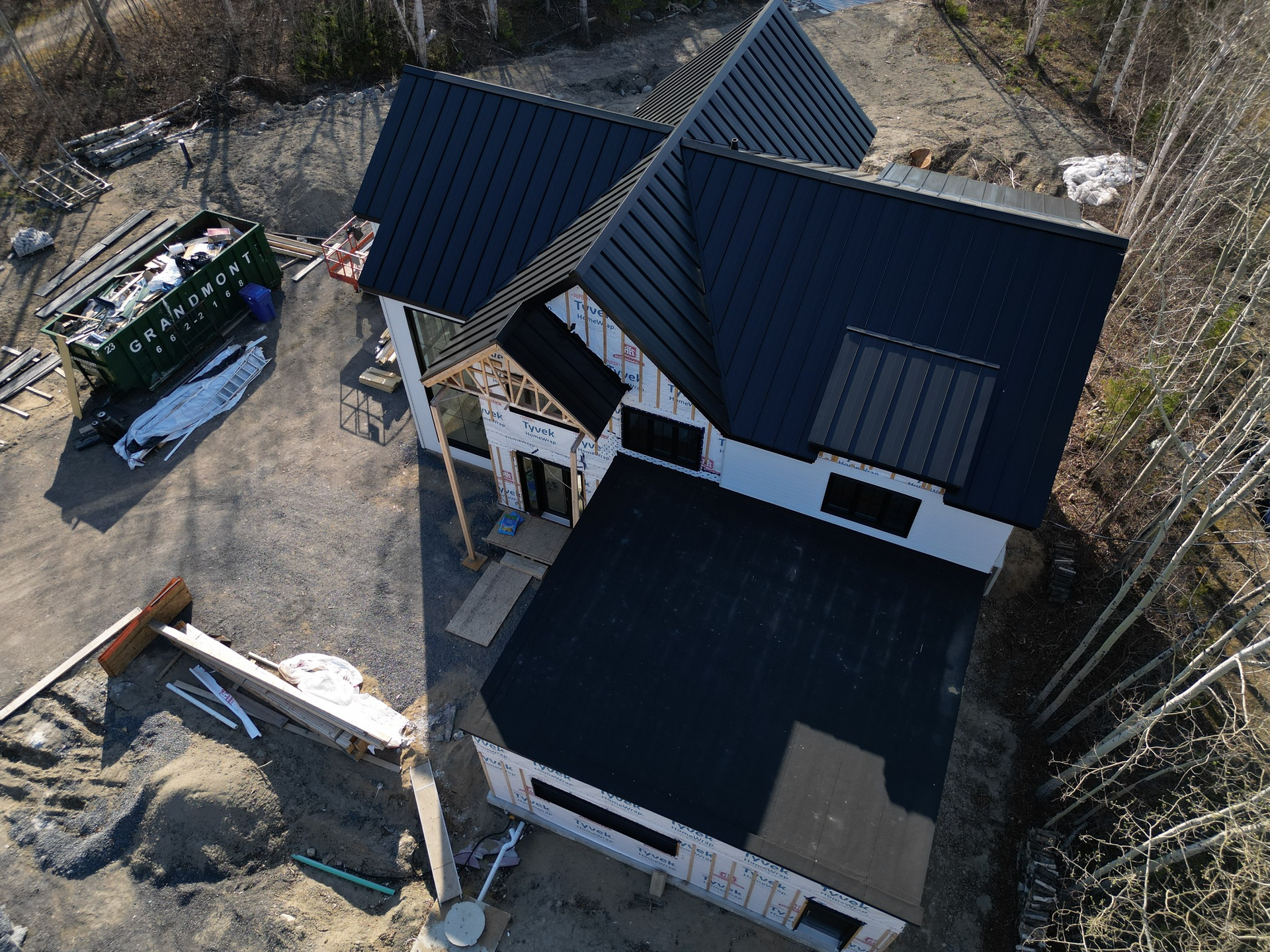 Vue aérienne d'une maison en construction, avec un toit sombre et un bardage blanc ; des débris de construction sont visibles.