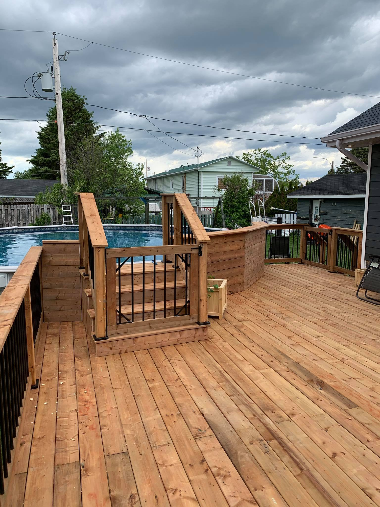 Terrasse en bois avec marches menant à une piscine. Rambarde noire et ciel gris.