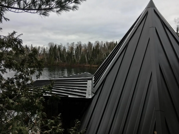 Toit en métal noir à pignon pointu, surplombant un lac entouré d'arbres sous un ciel nuageux.