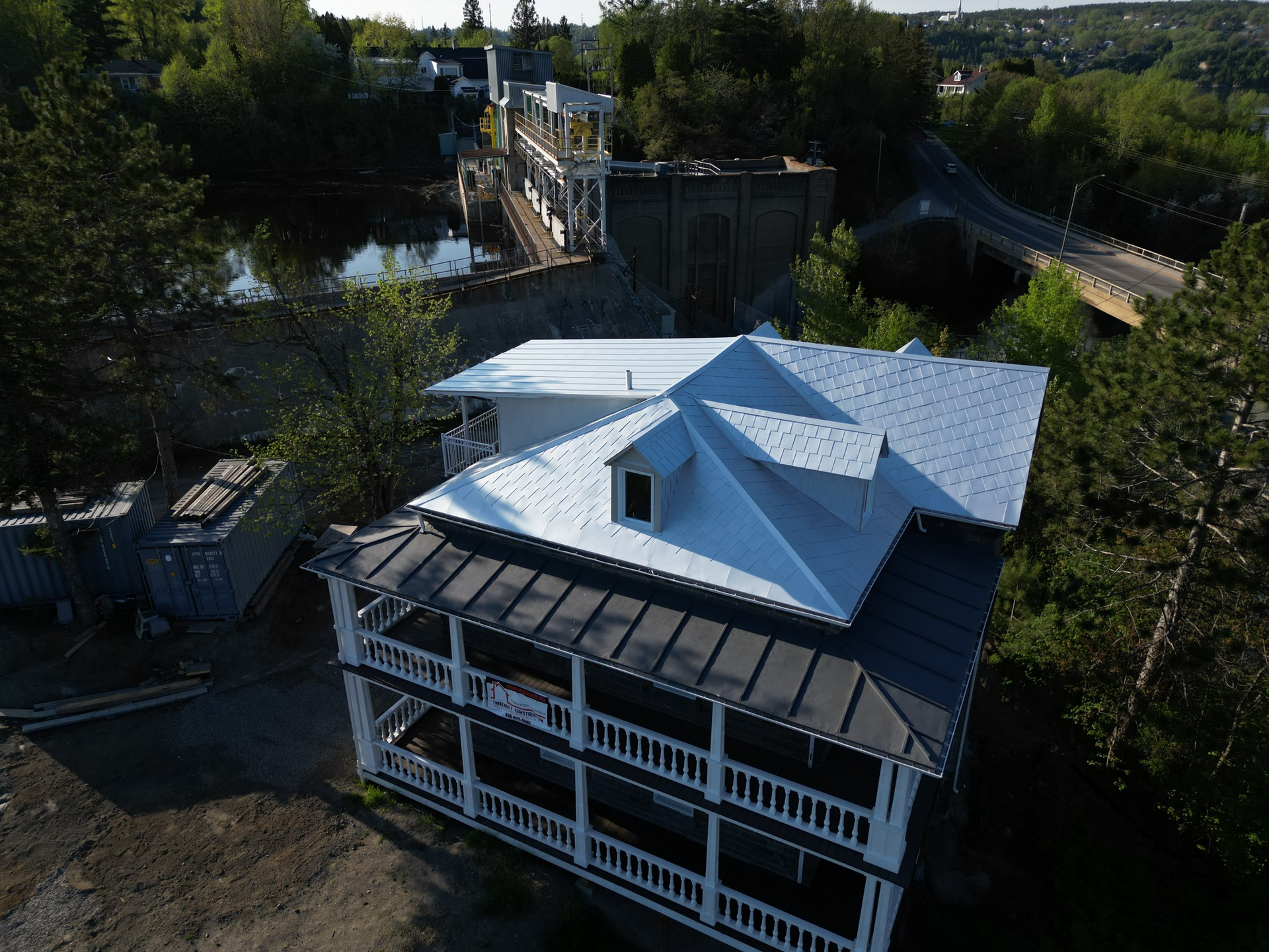 Bâtiment blanc de deux étages avec un toit noir et une véranda qui l'entoure, près d'une rivière et d'un pont.