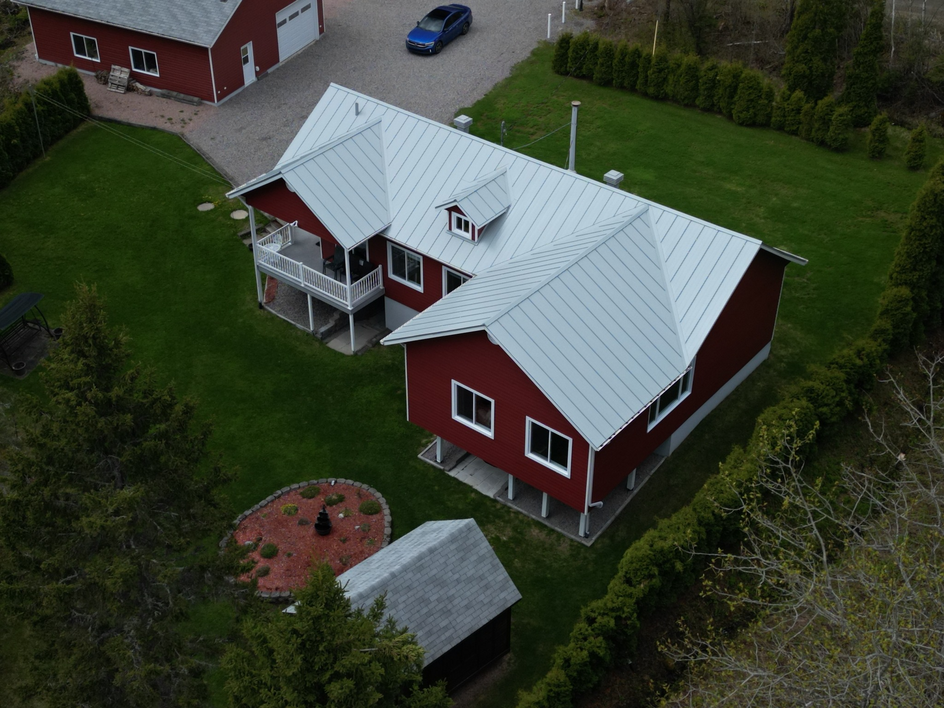 Maison rouge au toit argenté dans un jardin vert, avec un abri de jardin et une voiture.