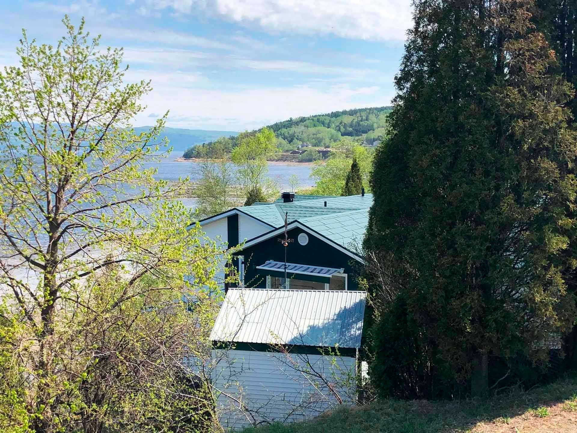 Une maison au toit turquoise et à l'extérieur blanc, encadrée par des arbres, surplombe un lac et son rivage lointain par une journée ensoleillée.