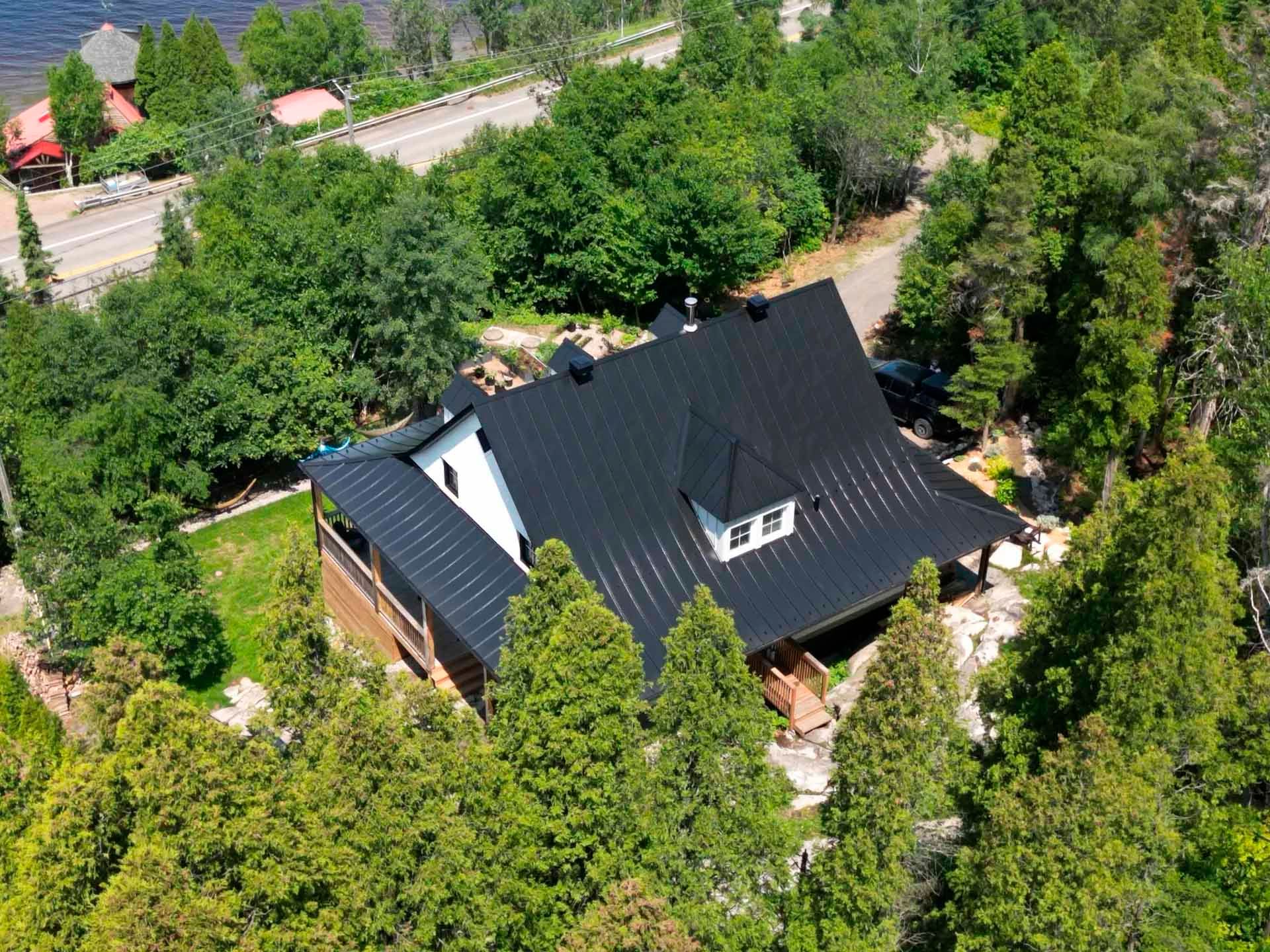 Vue aérienne d'une maison au toit en métal noir, entourée d'arbres, près d'une route et d'un lac.