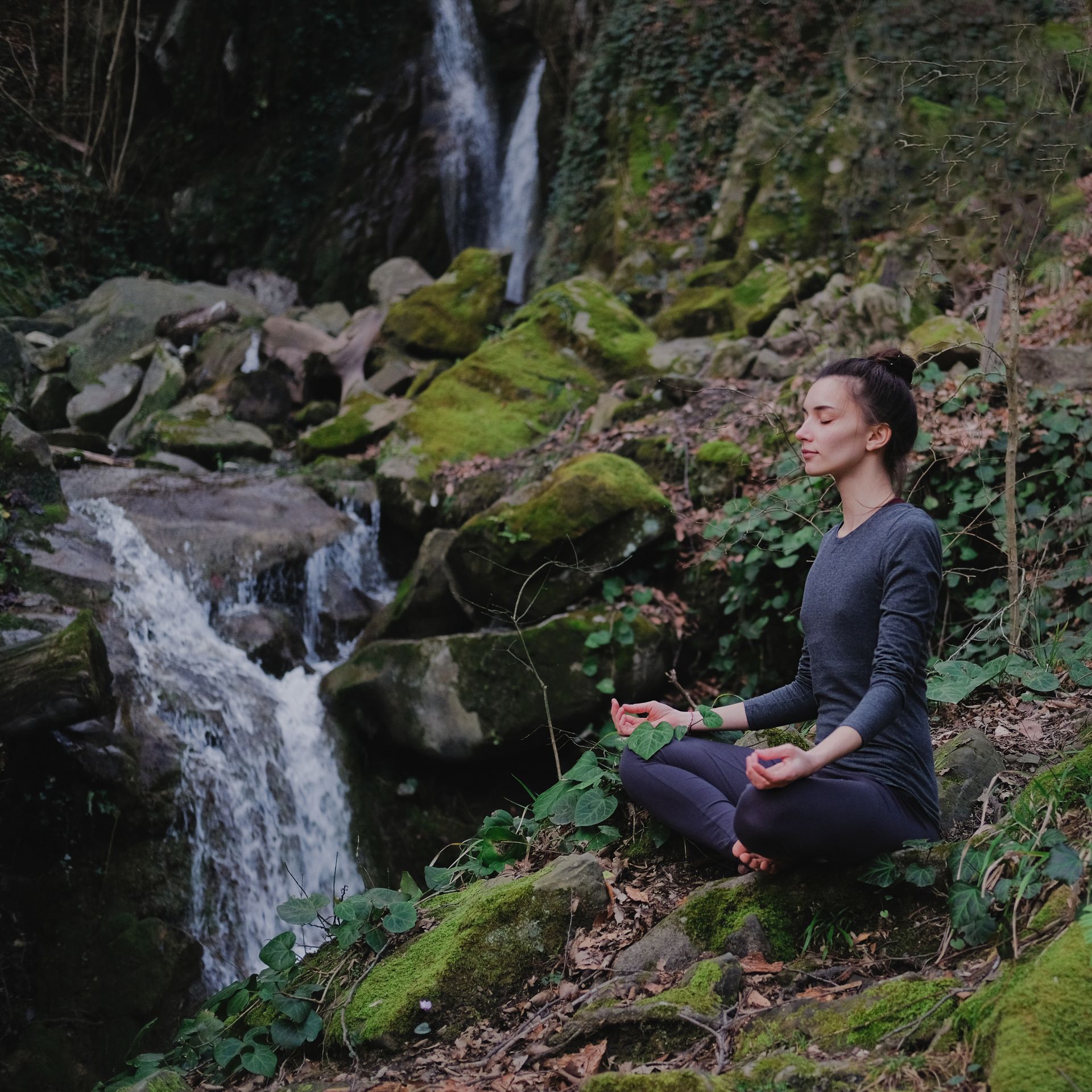 A person sits in a meditative pose on a mossy rock beside a cascading forest waterfall.