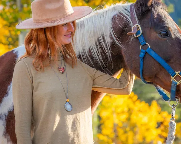 A person wearing a tan hat and shirt pets a brown and white horse in front of golden autumn trees.