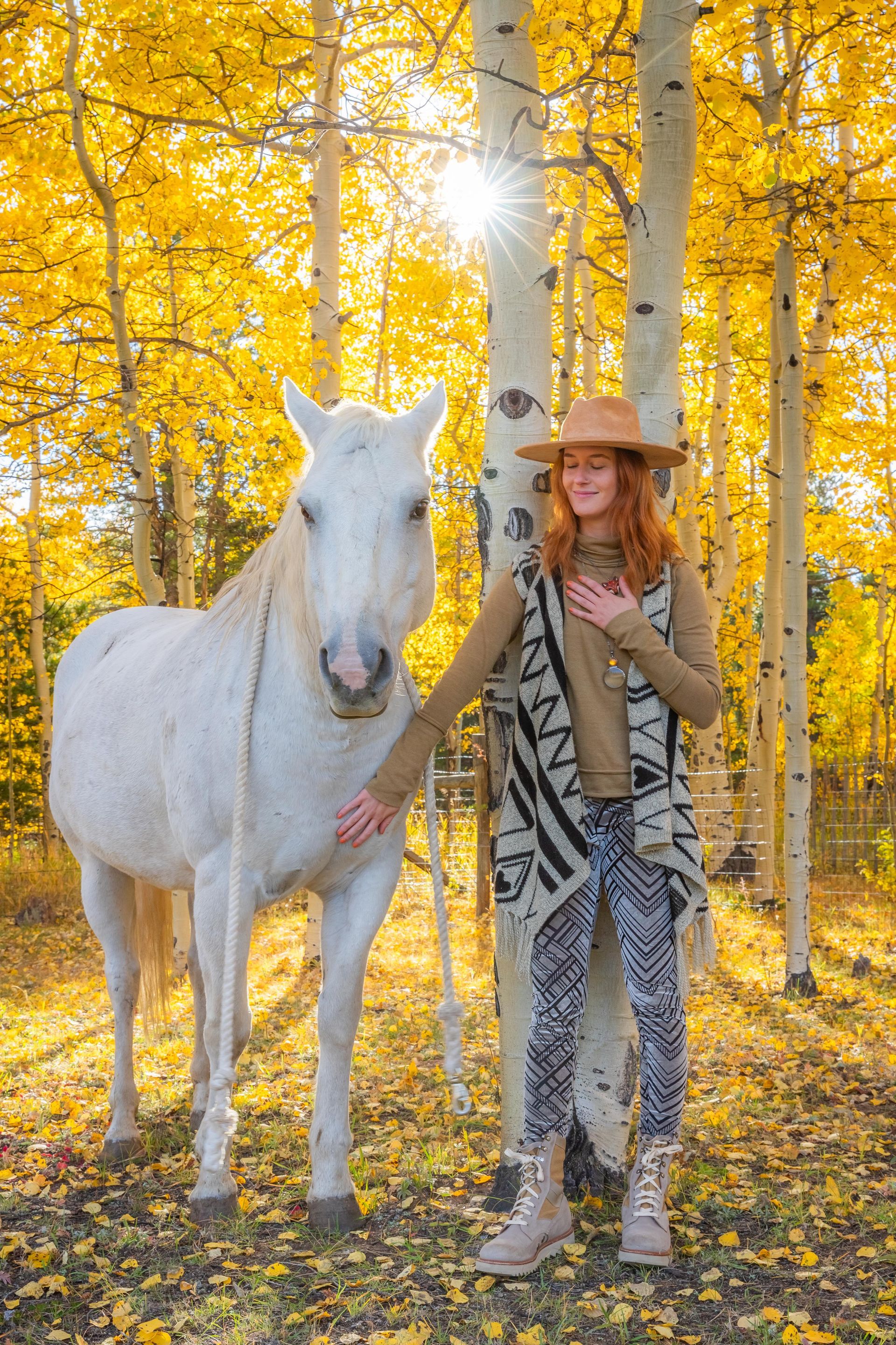 A person in a hat and patterned vest stands with a white horse in an autumn forest with bright yellow aspen trees.