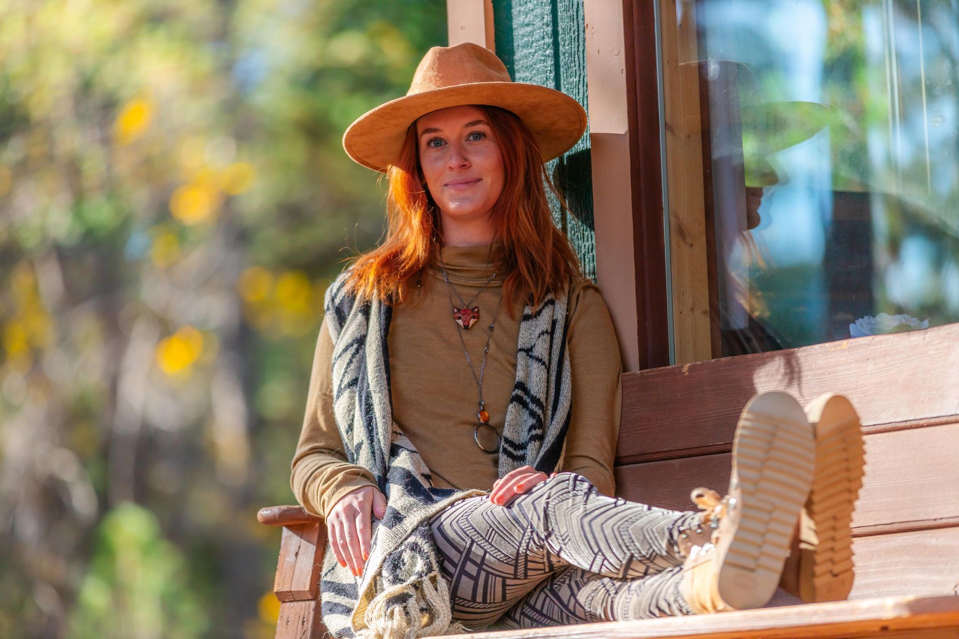 A person with red hair wearing a tan hat and sweater sits on a wooden bench outdoors, legs propped up, looking at camera.