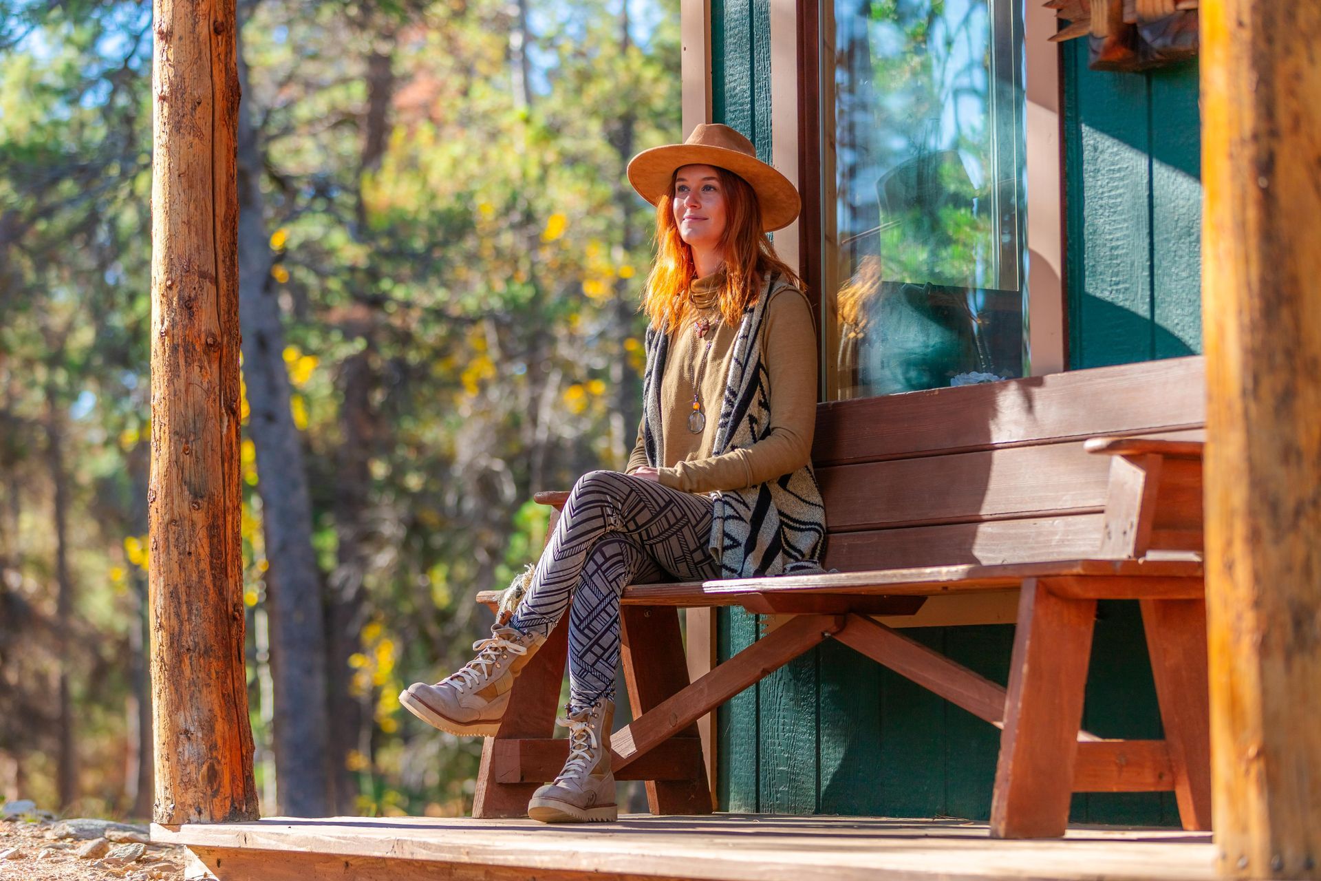 A woman wearing a hat and patterned vest sits on a rustic wooden bench on a porch, overlooking a sunlit forest.