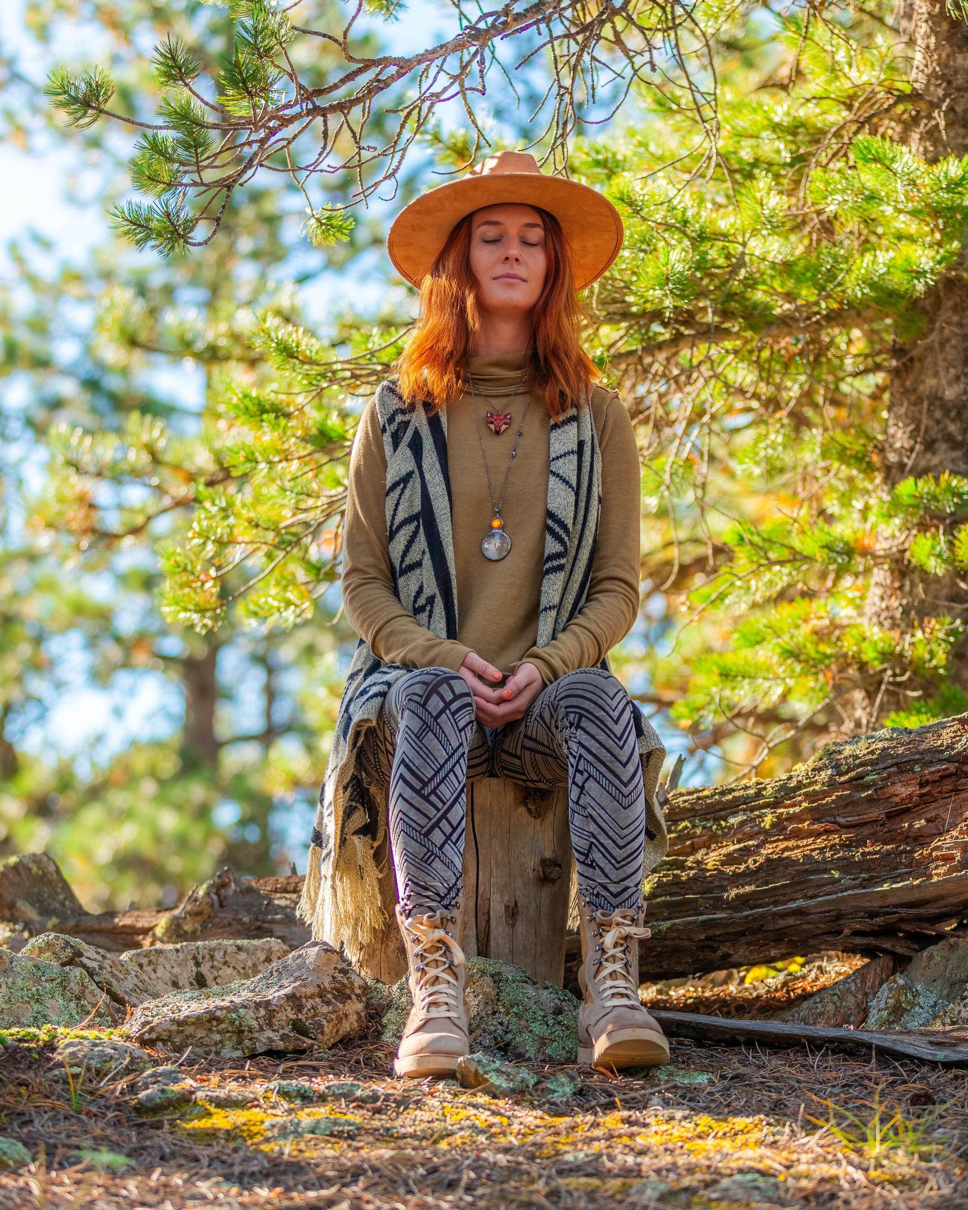 A person wearing a wide-brimmed hat, patterned vest, and leggings sits with eyes closed on a log in a sunlit forest.