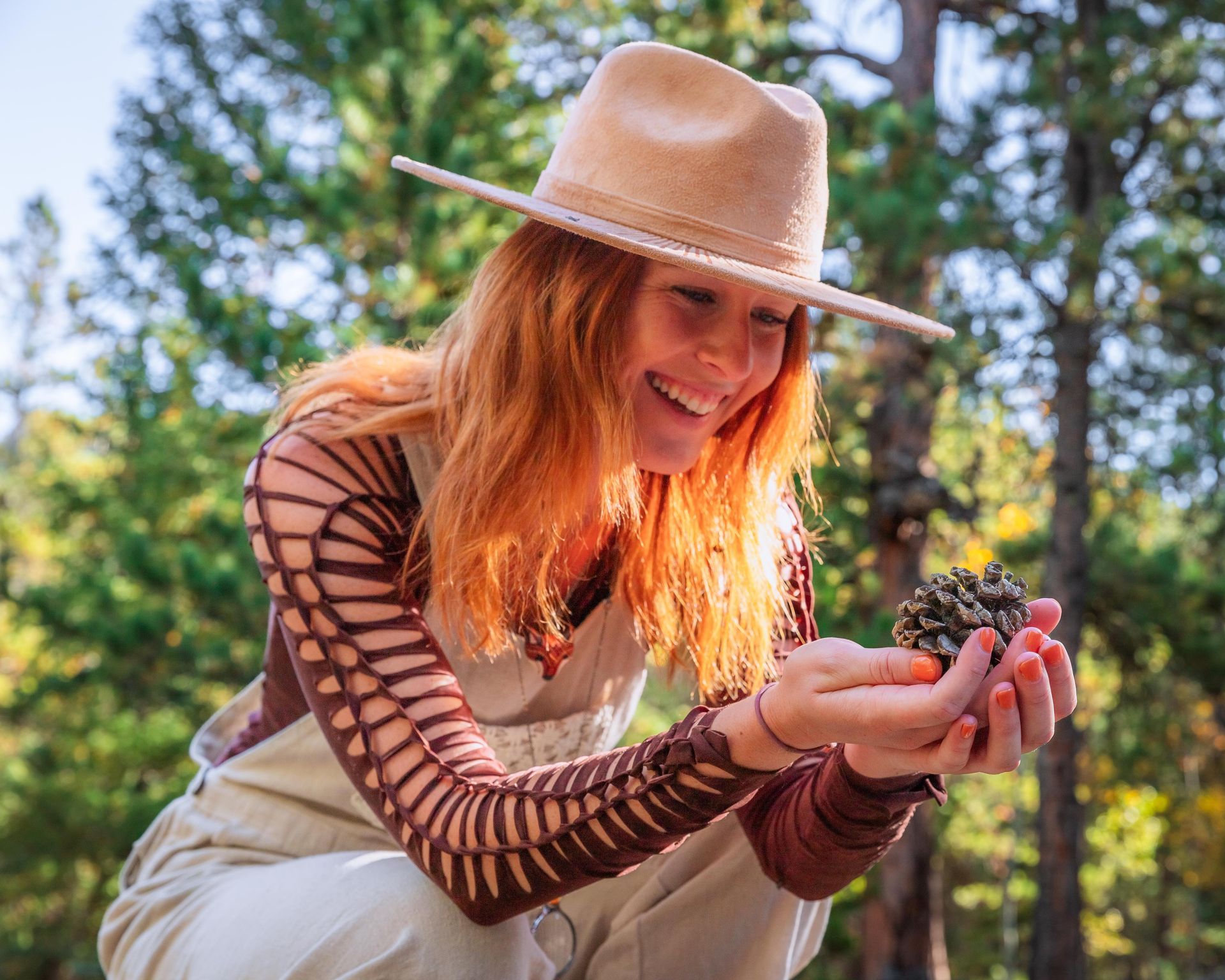 A person with reddish hair wearing a hat and patterned clothes sits on a wooden bench outdoors in a sunlit area.
