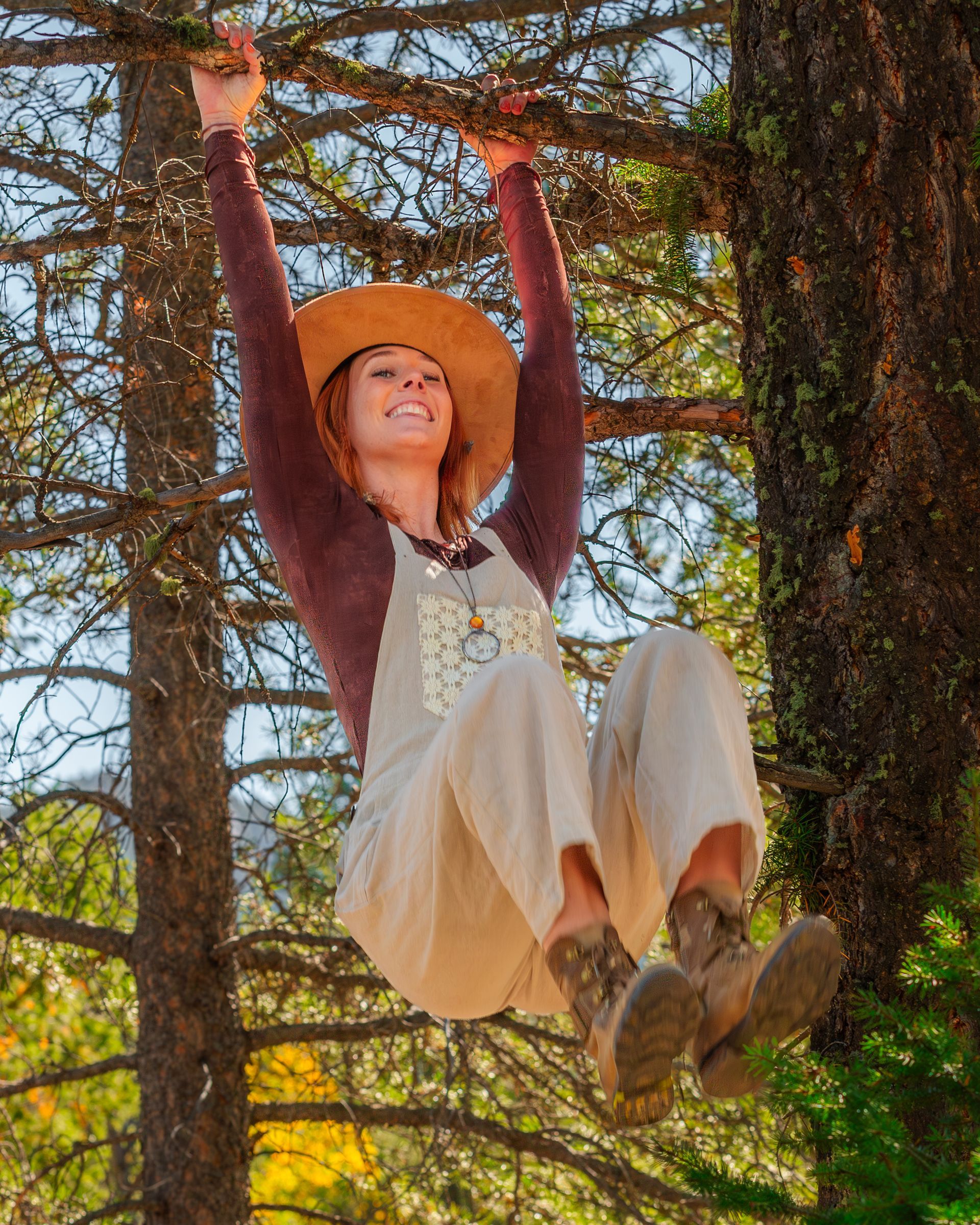 A person wearing a sun hat and overalls smiles while hanging from a tree branch in a sunlit forest.