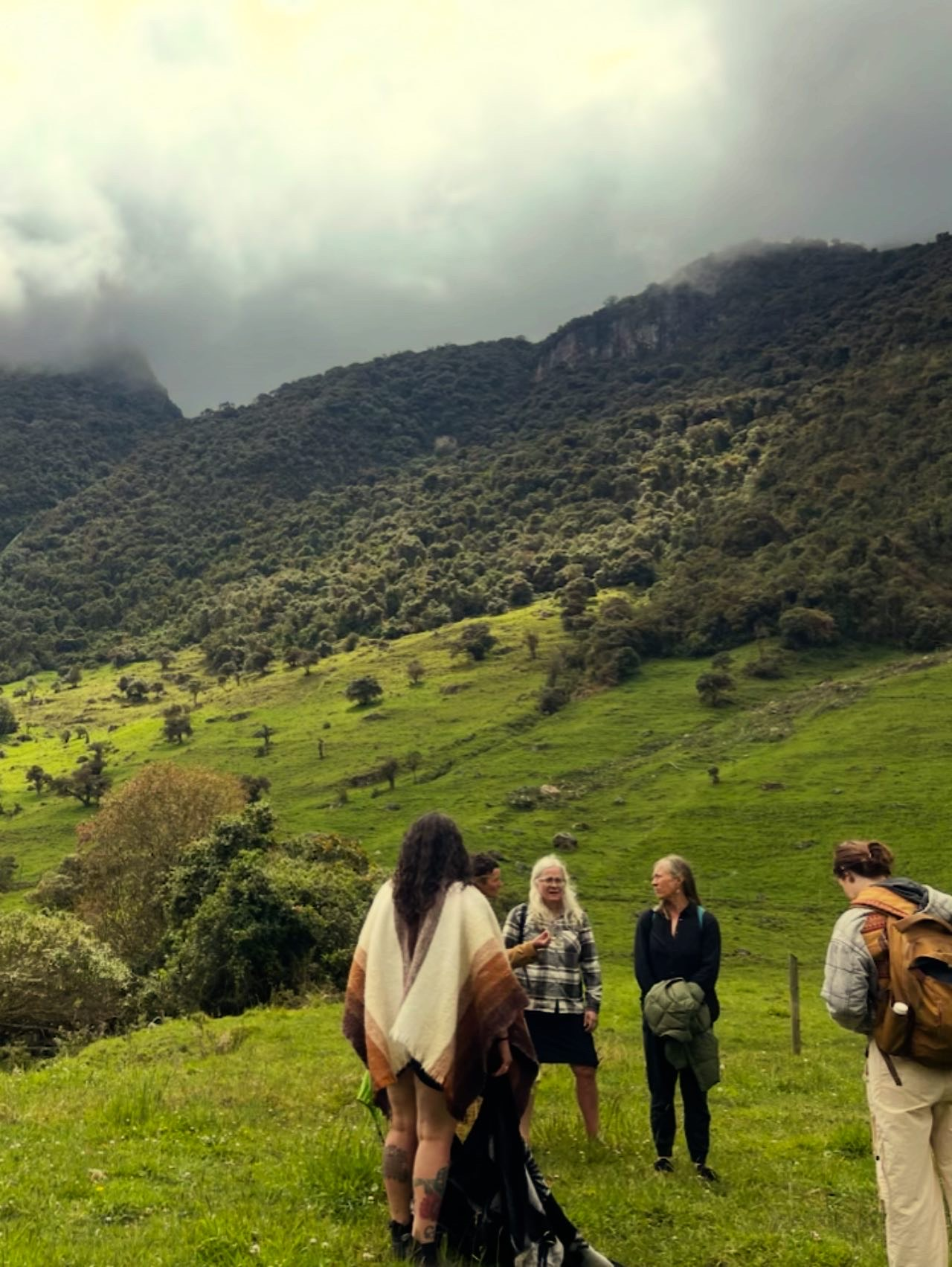 A person with reddish hair wearing a hat and patterned clothes sits on a wooden bench outdoors in a sunlit area.
