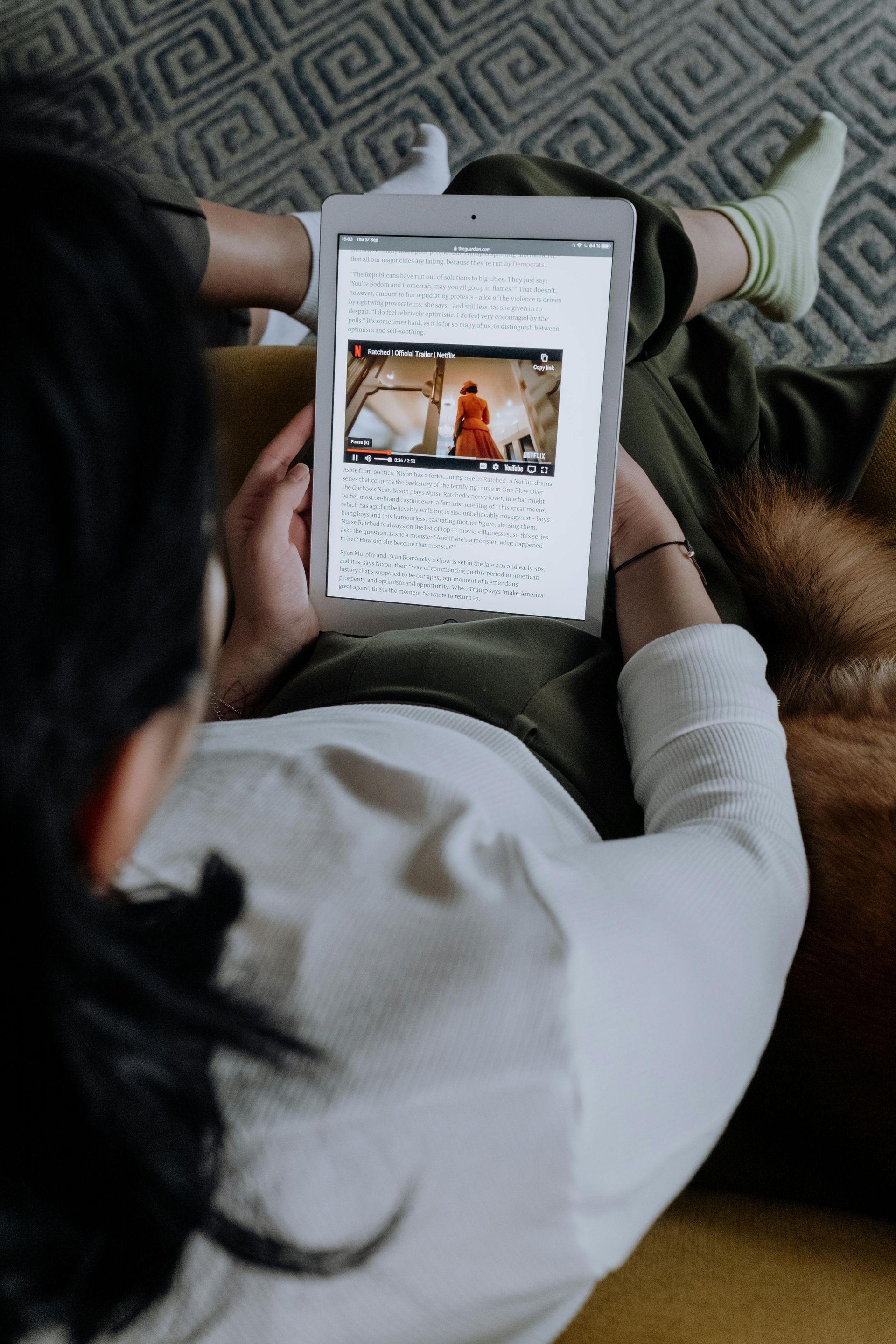 A person sitting on a patterned sofa, viewing a video on a tablet held in their hands.