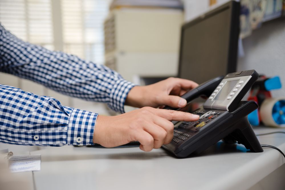 Hands Using a Black Office Phone on A Desk Beside a Keyboard and Monitor — Comms Cabling Central Coast In Kanwal, NSW