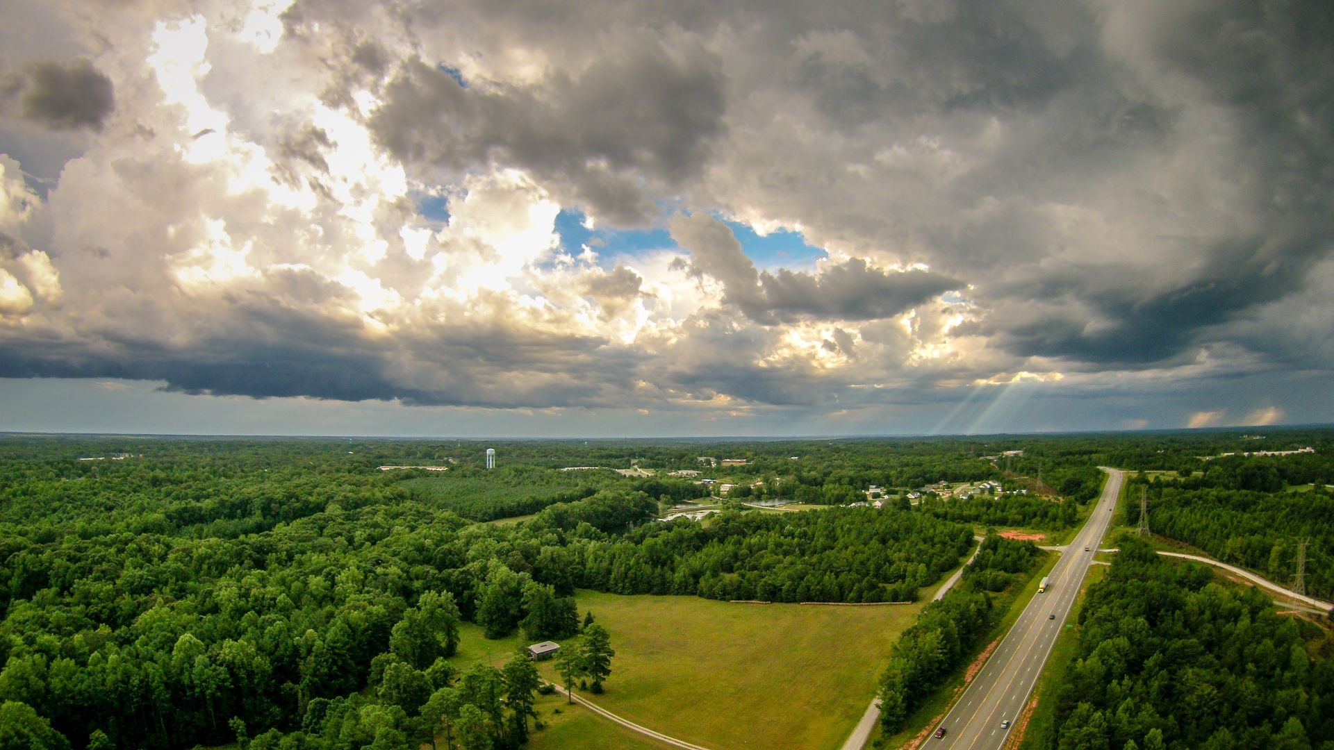 Overhead view of a green forest under a cloudy sky with sunlight piercing through. A highway is visible on the right.