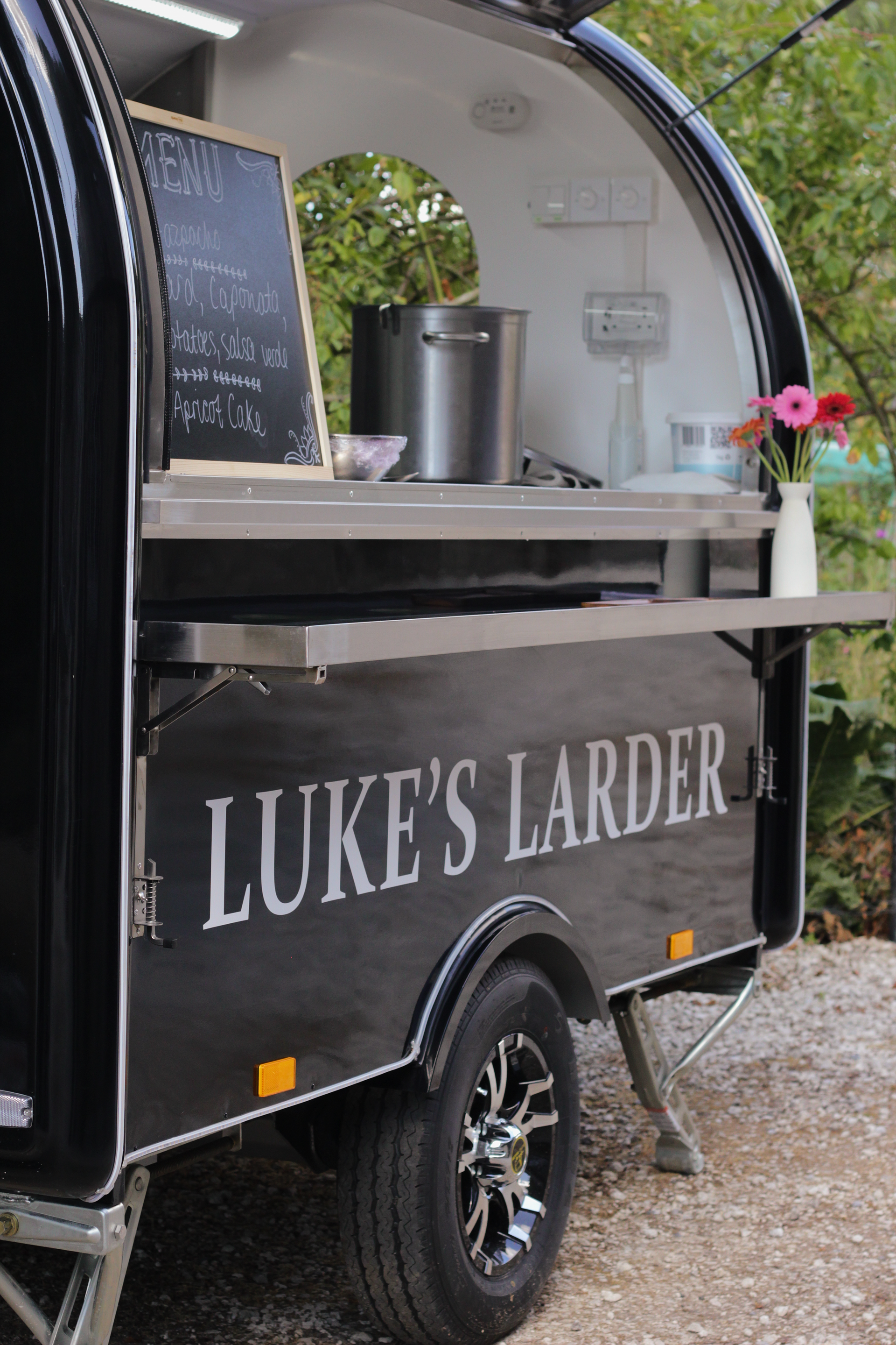 Luke’s Larder food trailer with serving counter, chalkboard menu, and potted flowers outdoors