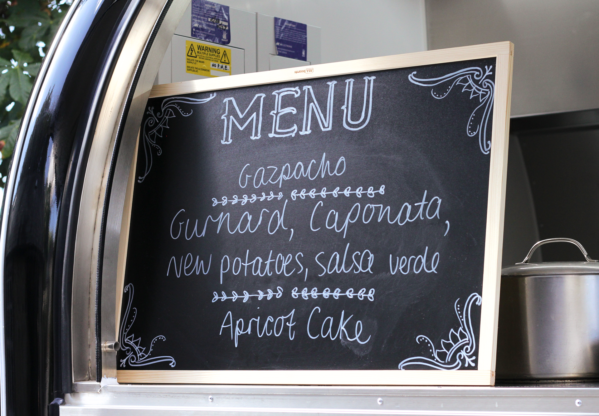 Chalkboard menu on a food truck counter listing gazpacho, salad, and apricot cake.