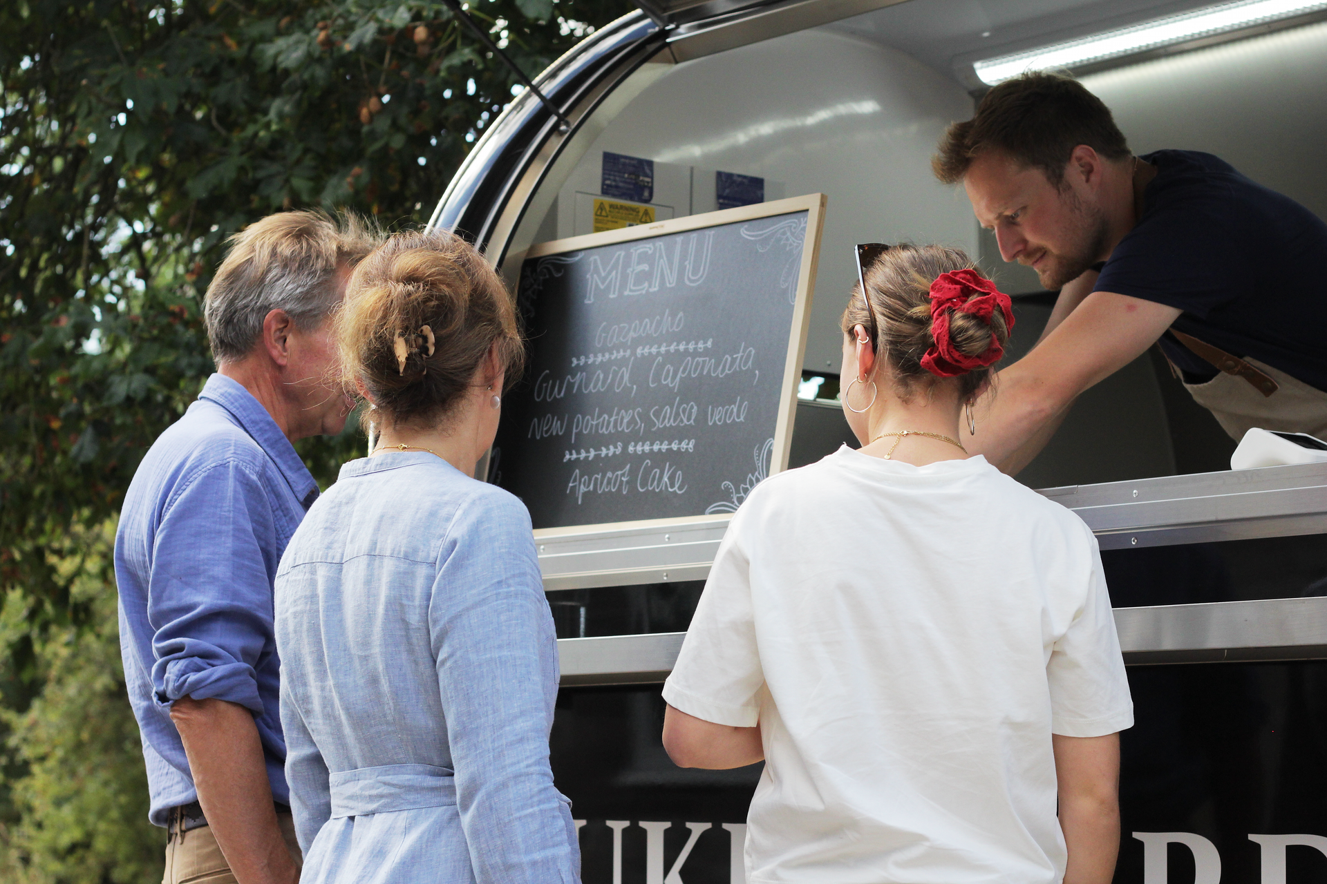 People gathered at a food truck as a worker writes on a chalkboard menu.