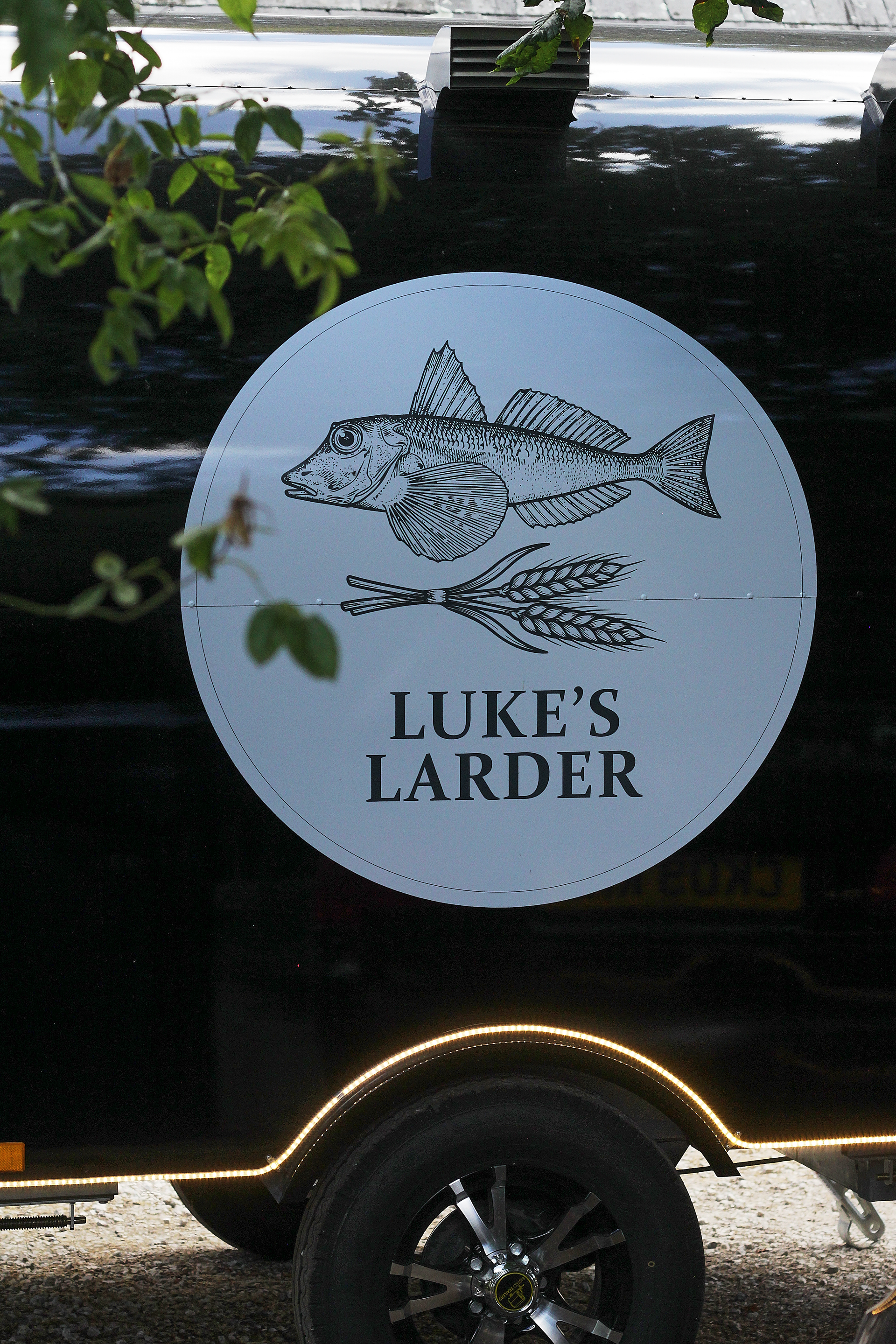 Luke’s Larder logo with fish on a white circle on a black vehicle side, with tree branches above.