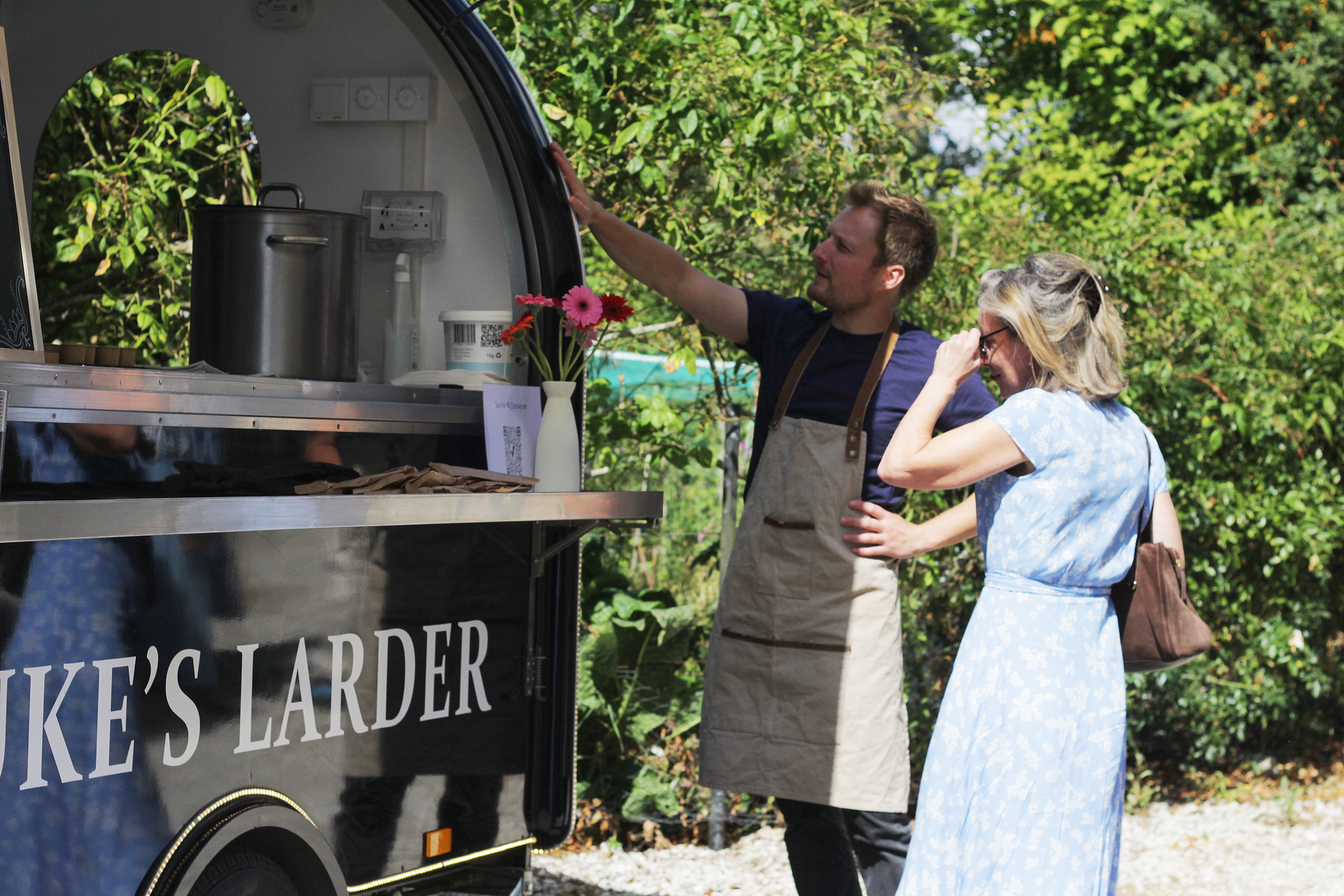 Two people at an outdoor coffee cart, one pointing up while the other looks on with a hand on her hip.