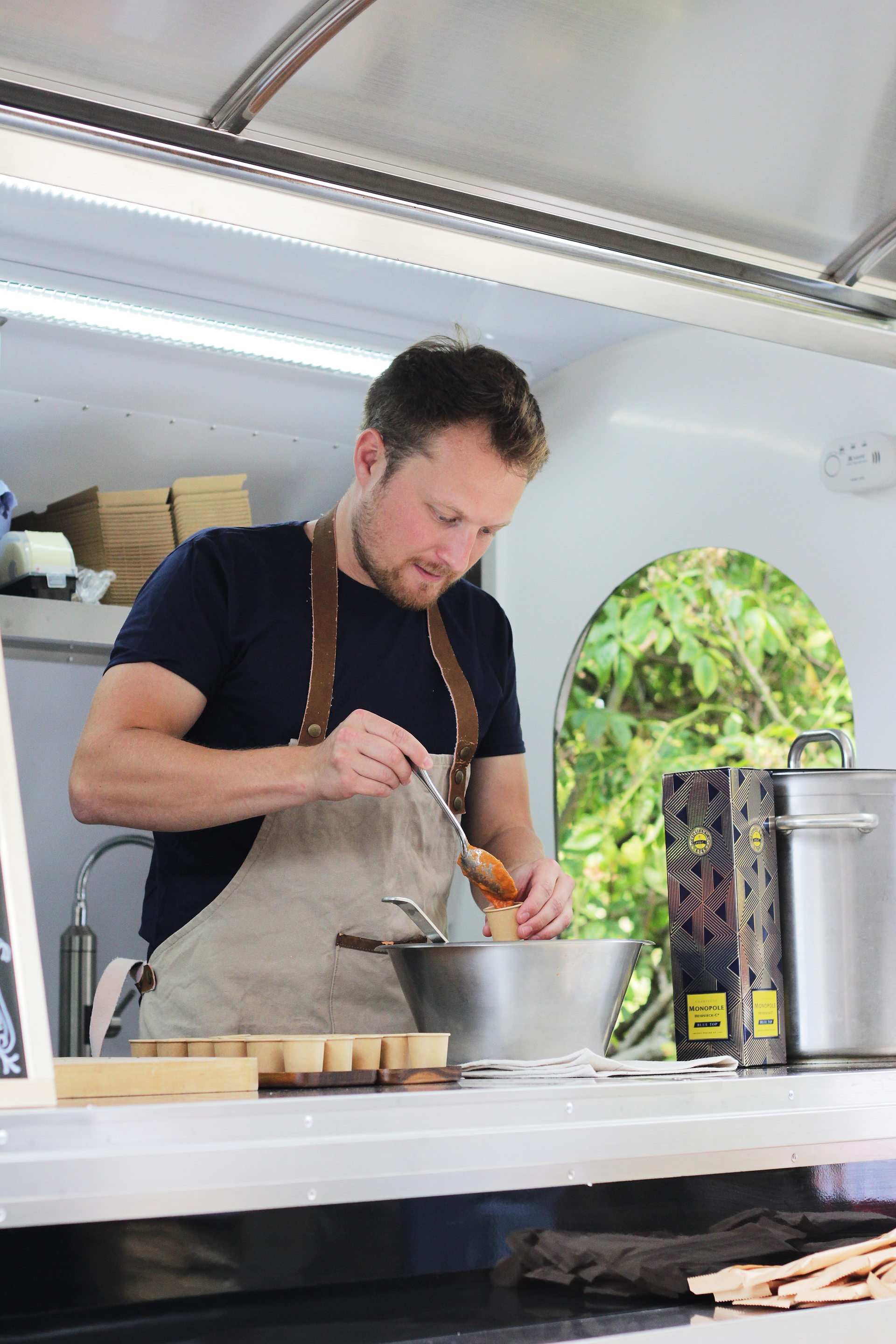 Man in a navy shirt preparing food at an outdoor market stall, stirring a pot beside stacked containers.