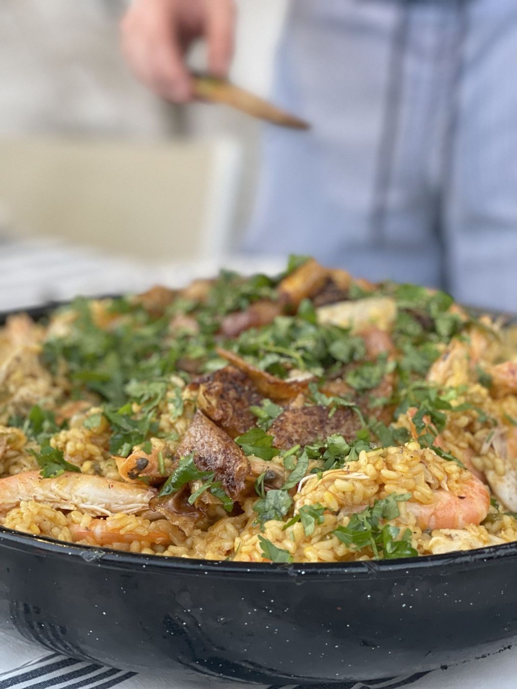 A skillet of saucy food topped with herbs, with a person serving in the background.