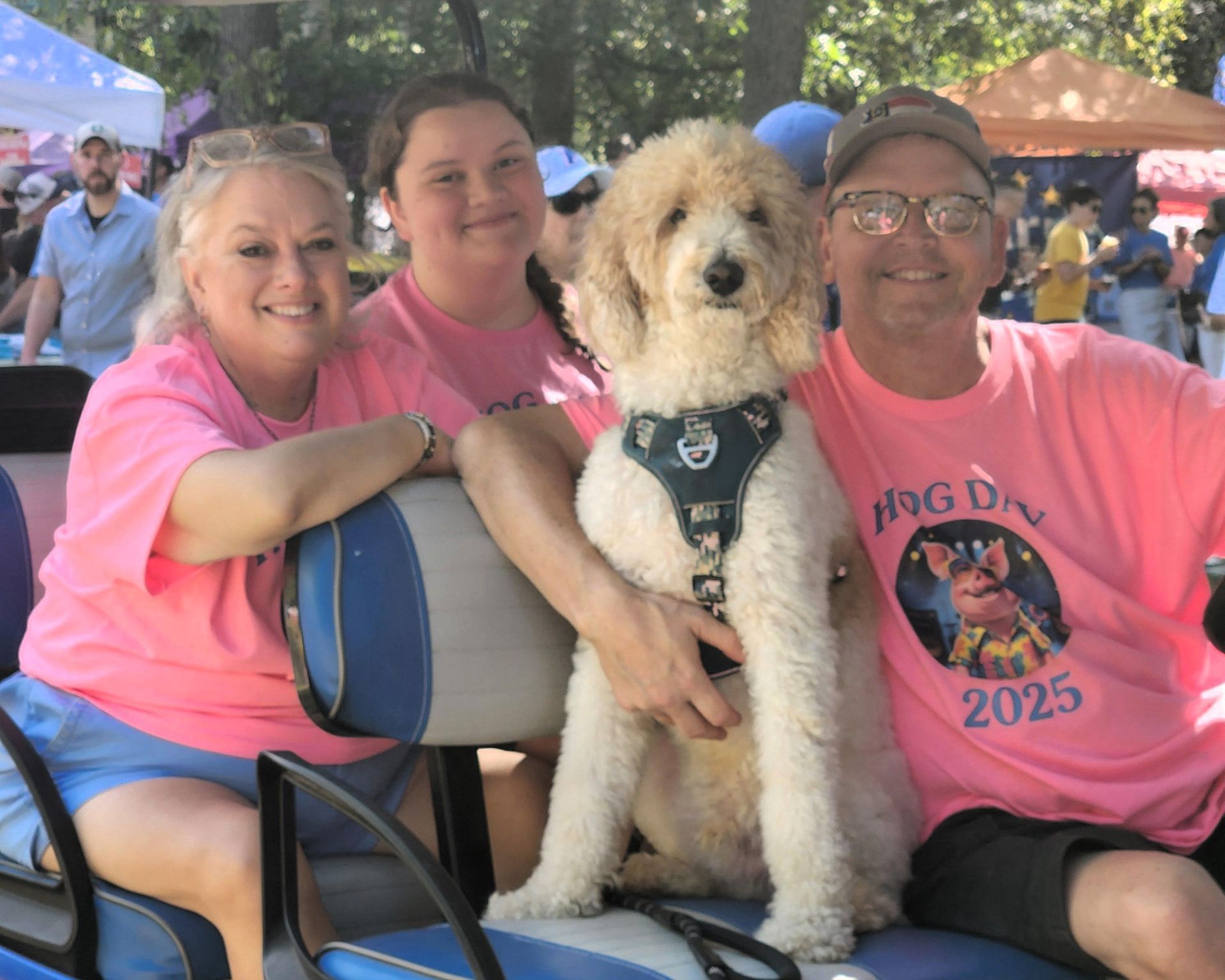 Three people with a fluffy dog at an outdoor event, all wearing pink shirts, seated at a table.