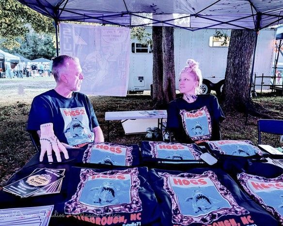 Two people in black graphic tees sit at a table under a canopy, displaying printed shirts outdoors.