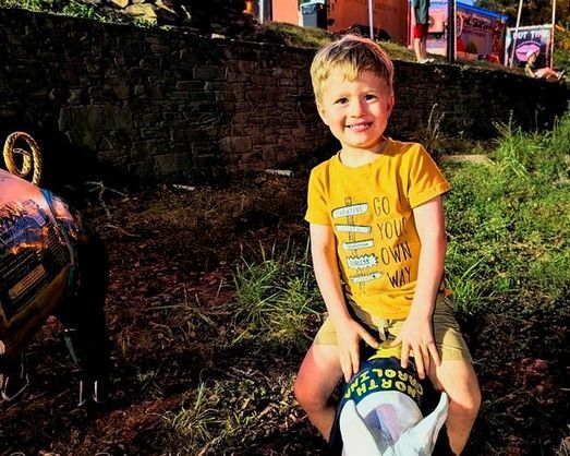 Child in a yellow shirt smiling while sitting on a ride outdoors.