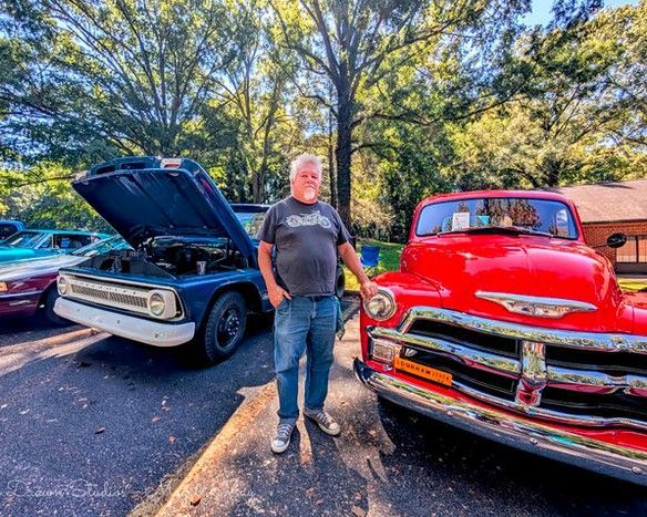 Man standing between a red classic truck and a black classic car with hoods open, in a sunny wooded lot