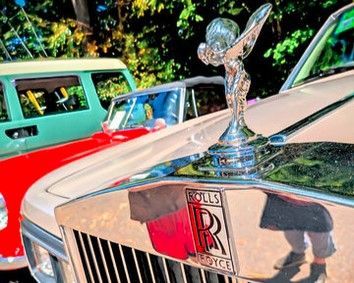 Close-up of a Rolls-Royce hood ornament and grille, with colorful classic cars in the background.