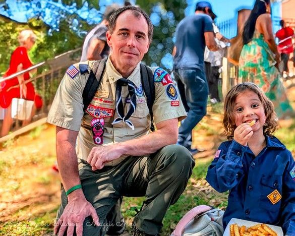 Two people crouch outdoors at a campsite, one wearing scout badges, with trees and people in the background.