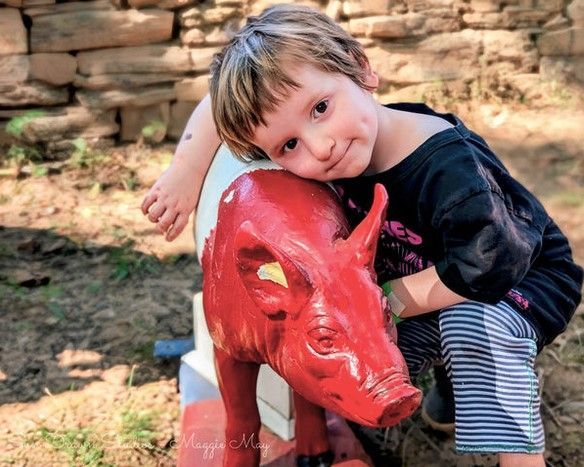 Child hugging a glossy red pig statue outdoors against a stone wall.