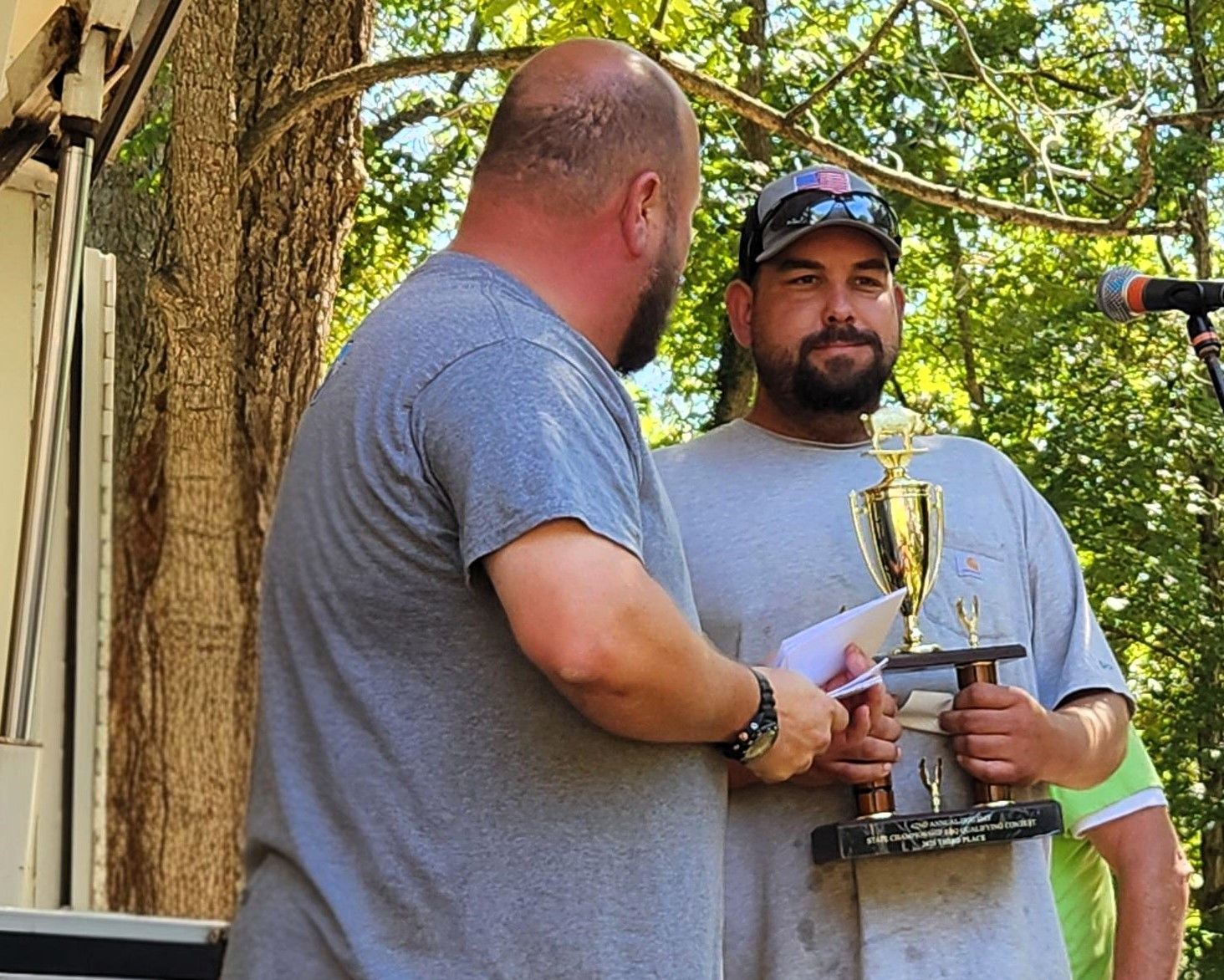 Two men on a stage exchanging a trophy outdoors under trees
