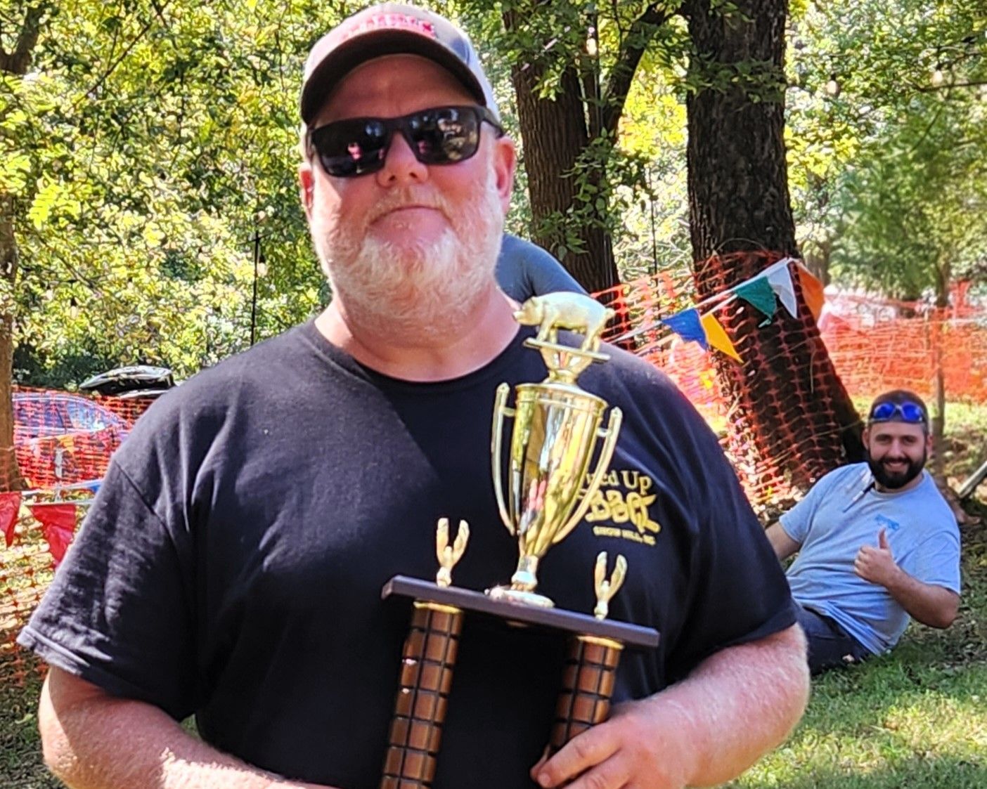 Man holding a trophy outdoors at a colorful event, with another man sitting in the background.