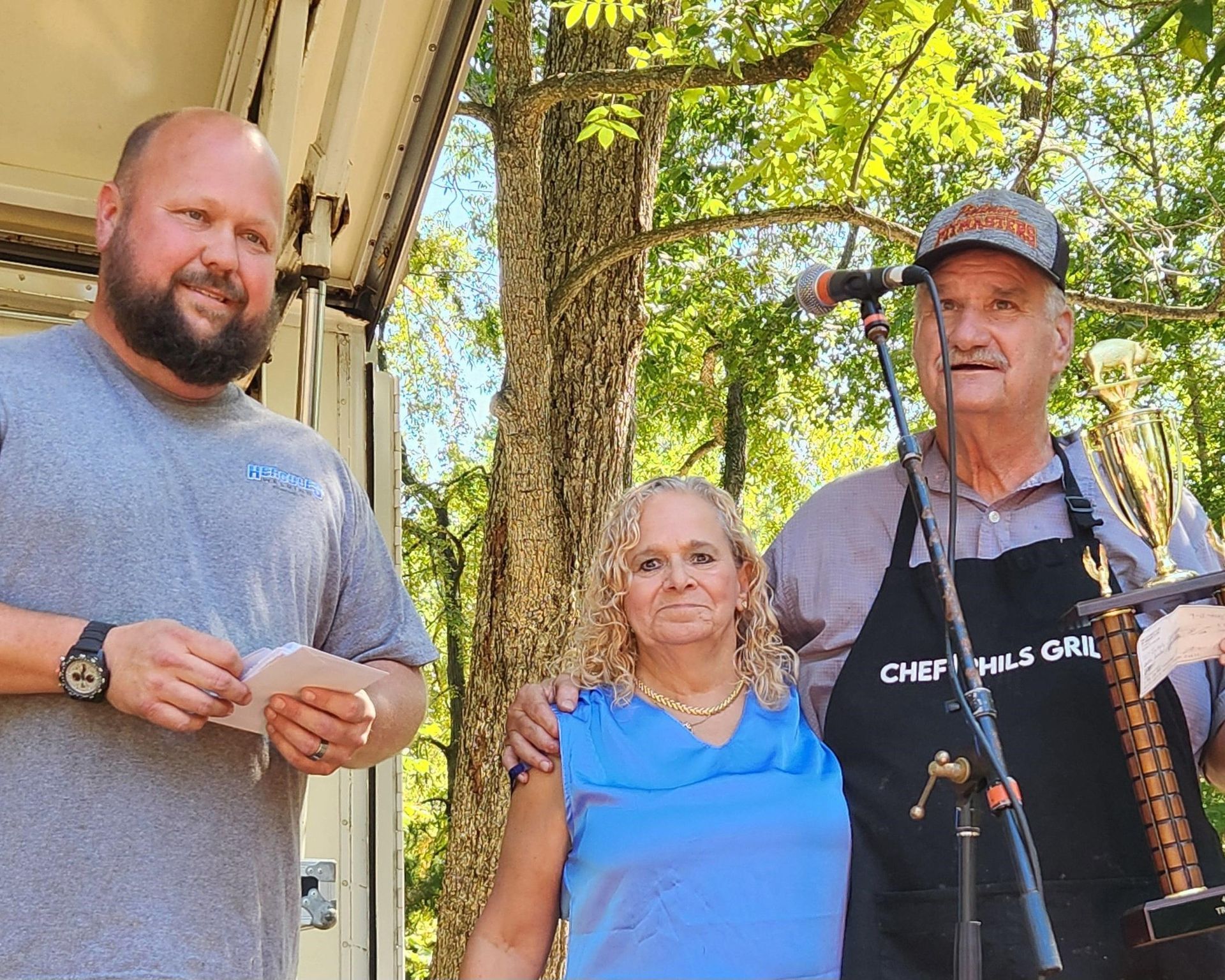 Three people standing outdoors near a microphone, one holding a stringed instrument, under trees.