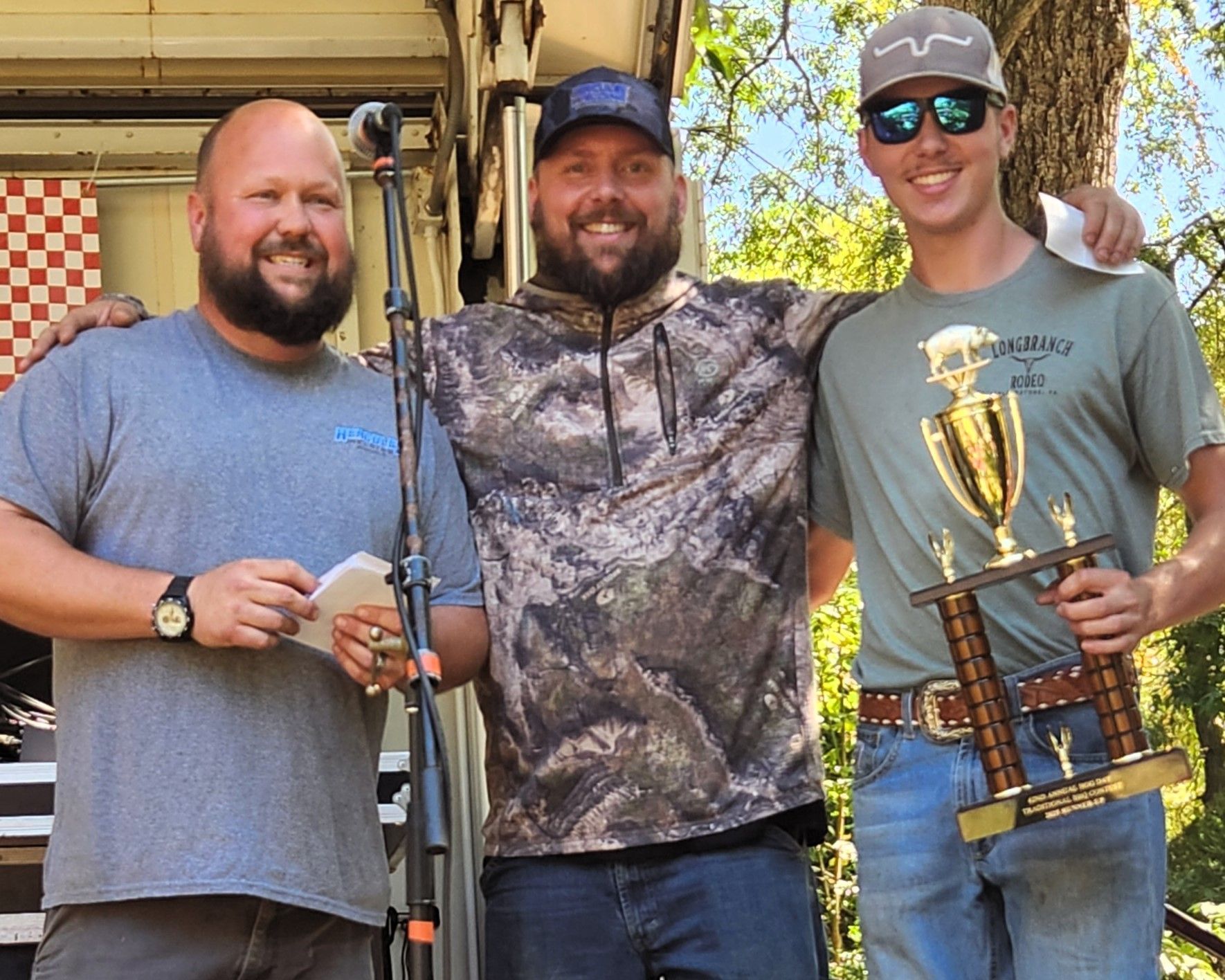 Three men smiling outdoors, one holding a large trophy and two holding long guns.