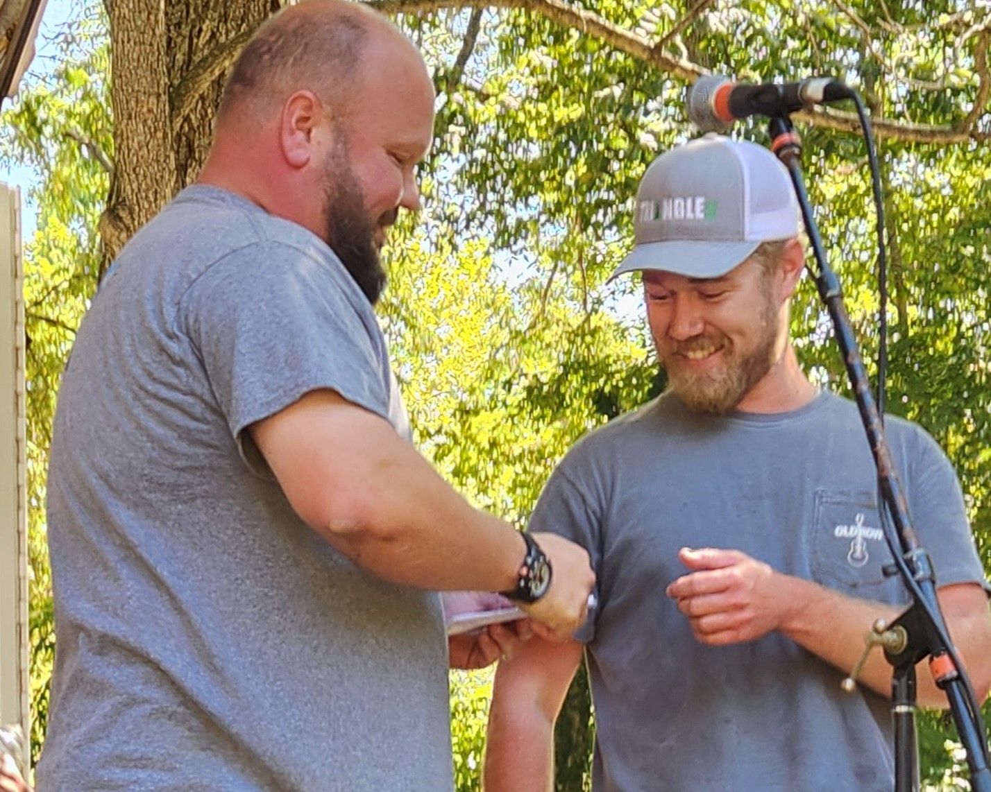 Two men in gray shirts smiling outdoors near a microphone stand, one checking the other’s wrist.