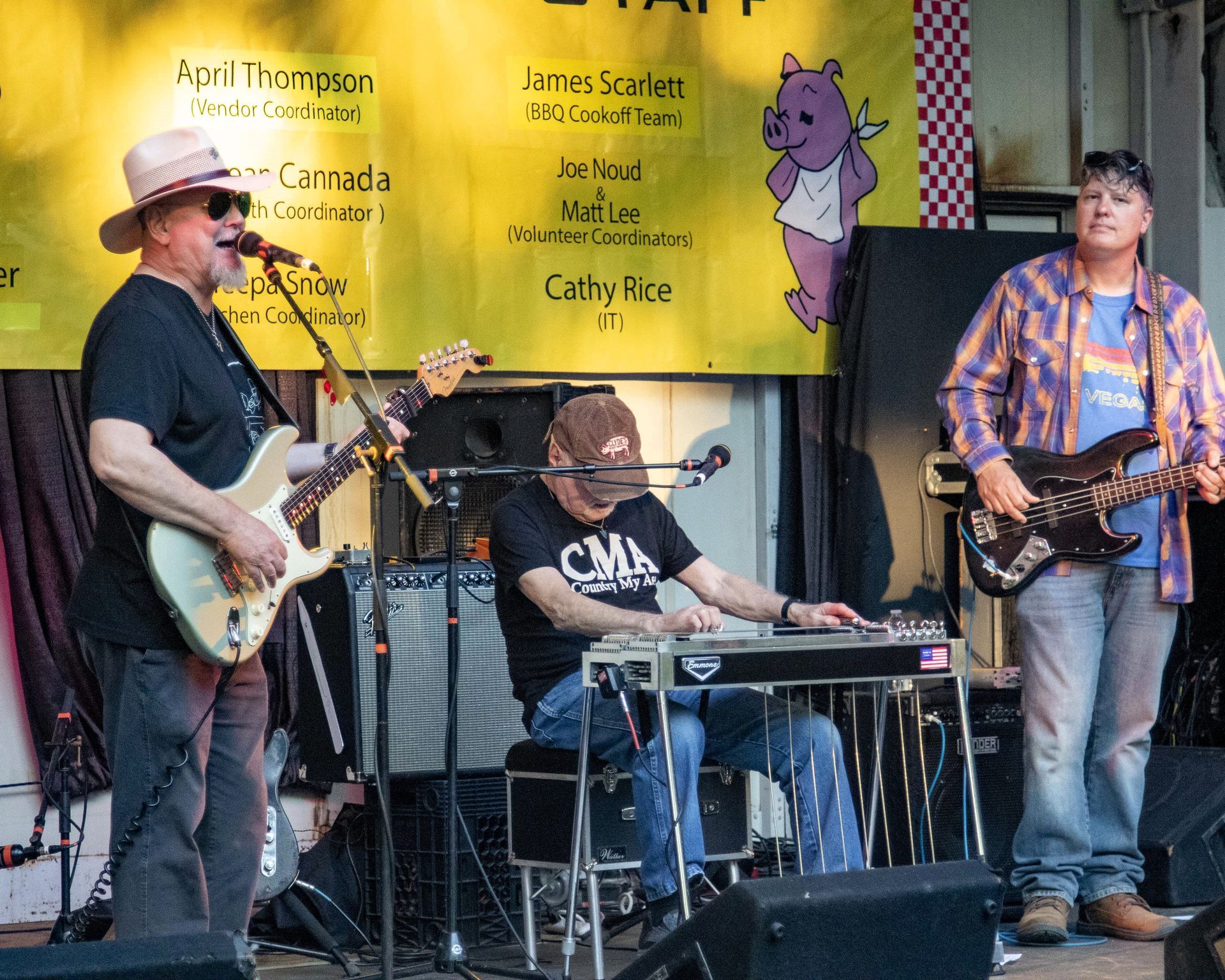 Band performing live on an outdoor stage with guitars, keyboard, and drums beneath a yellow banner.