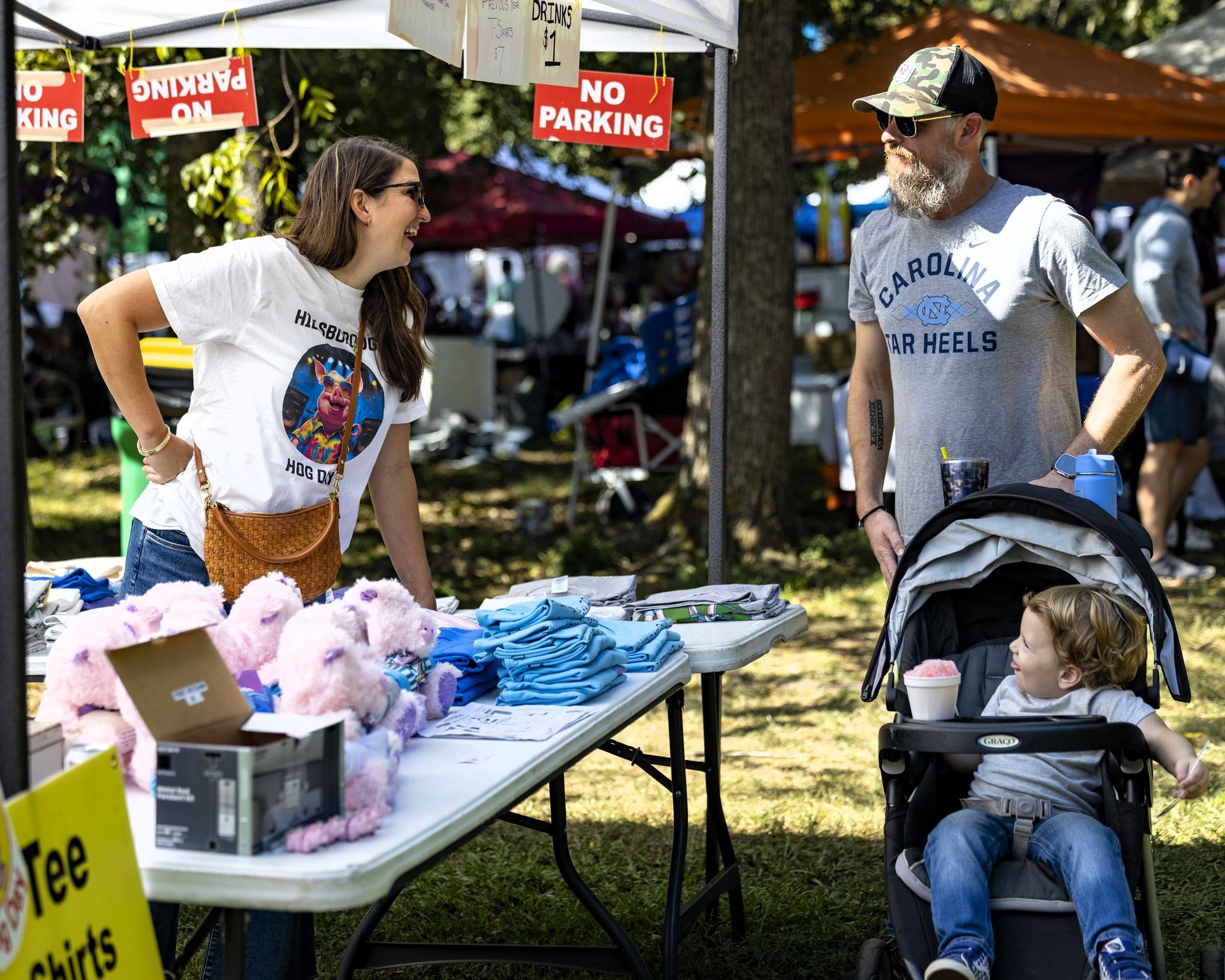 Vendor table with colorful shirts and plush toys at an outdoor market, with a man and child in a stroller nearby