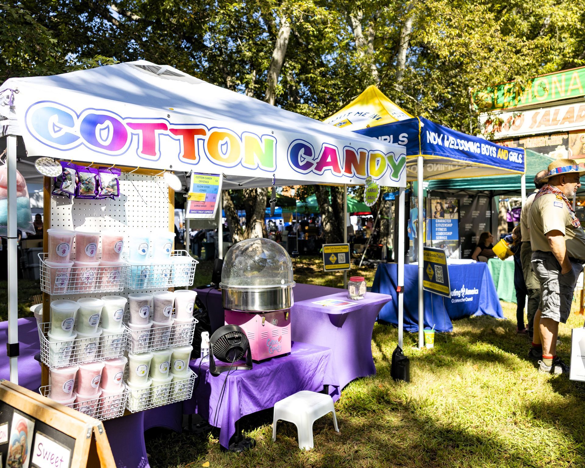 Cotton candy booth at an outdoor fair with colorful tents, stacked containers, and a vendor on the right