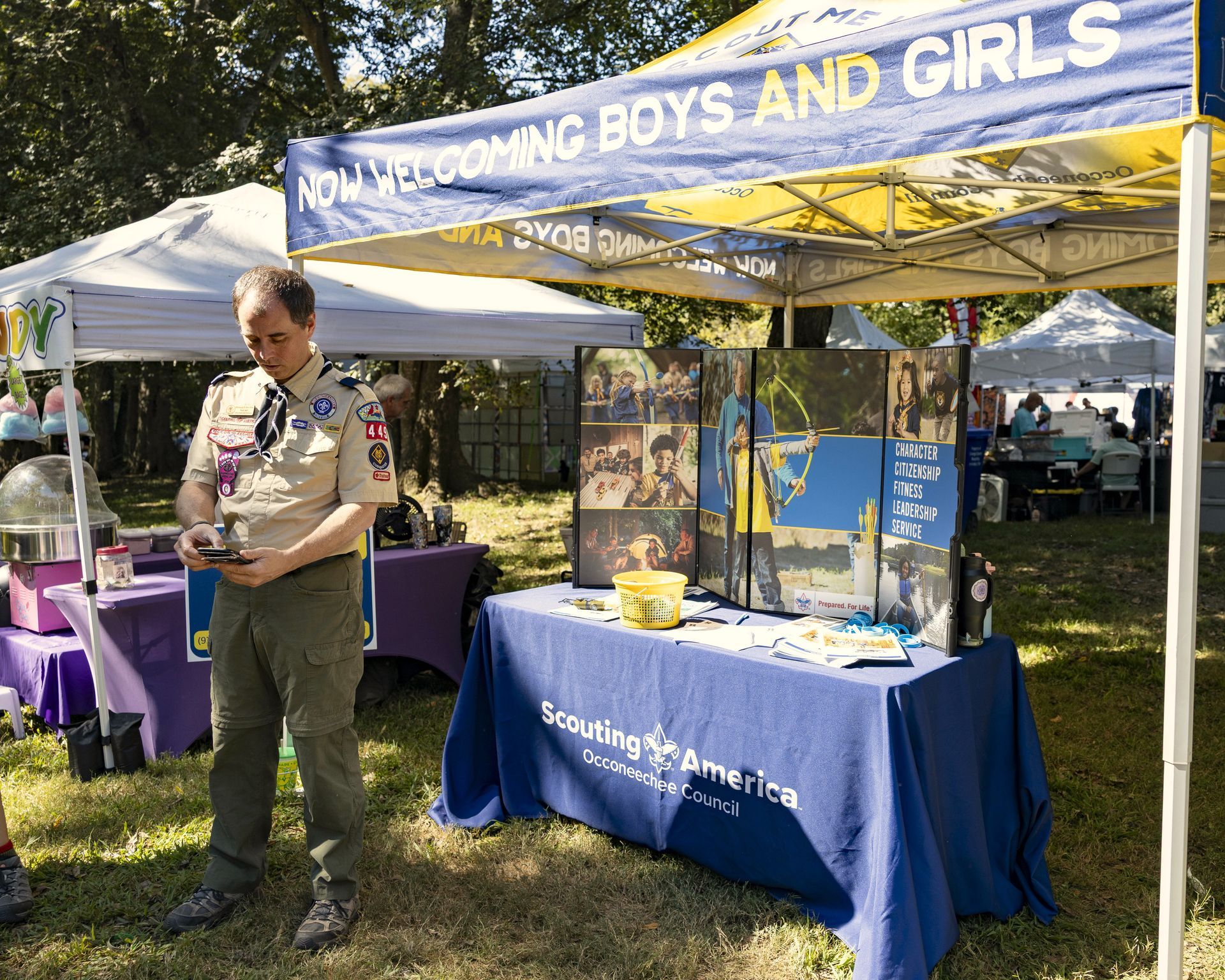 Outdoor booth with blue “BOYS AND GIRLS” banner, displays, and a man standing beside a purple table.