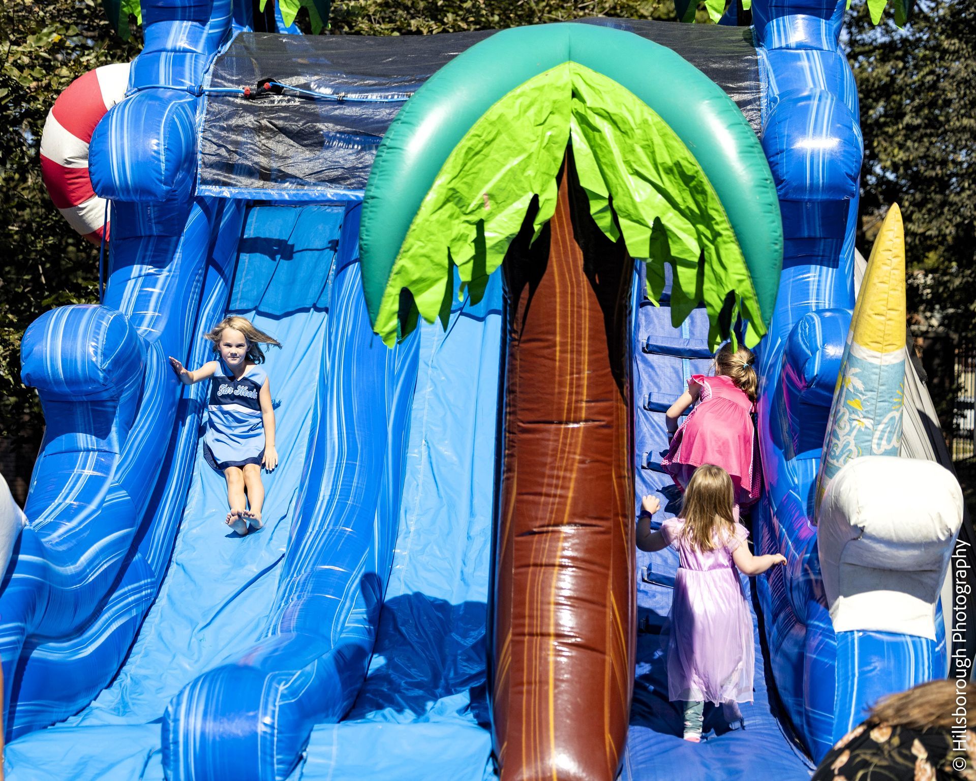 Children sliding on a blue inflatable water slide with a palm-tree centerpiece.