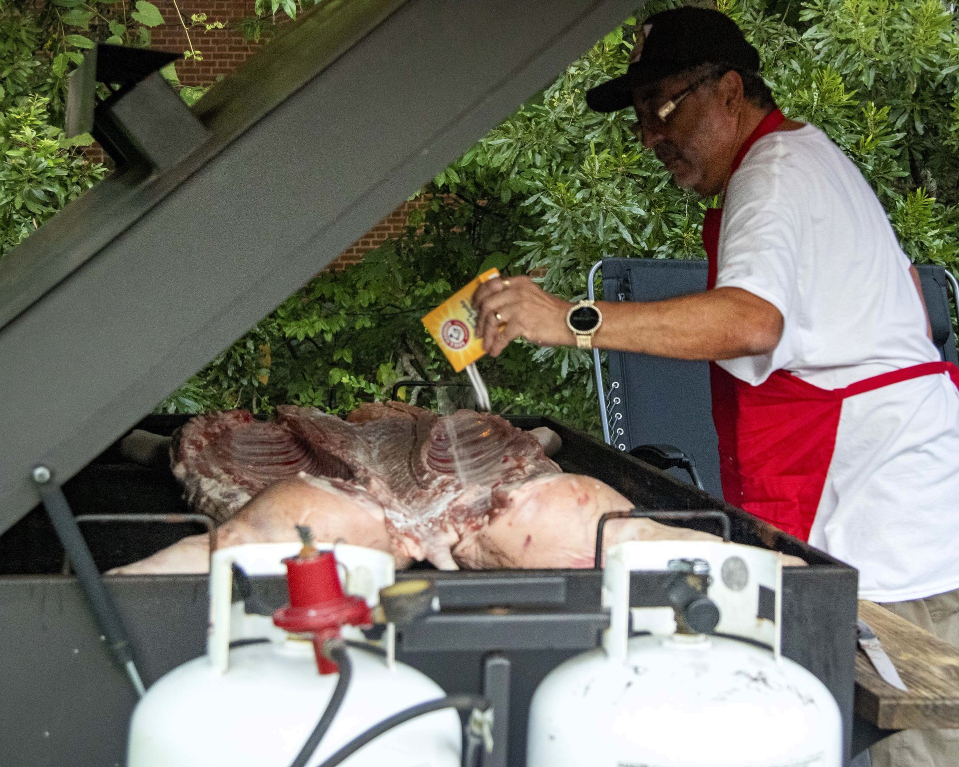 Man grilling a whole pig on a spit beside propane tanks outdoors