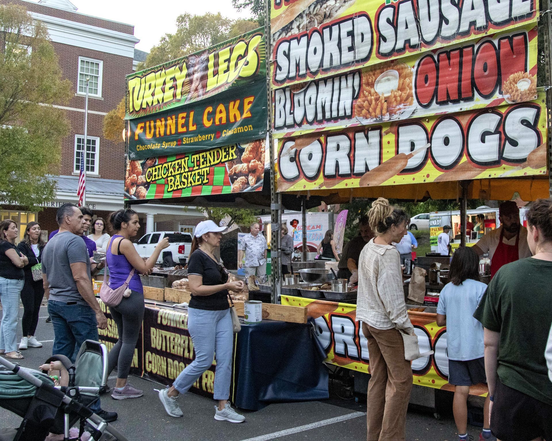 Crowded street food stalls with large signs for smoked sausage, brown corn dogs, and funnel cake